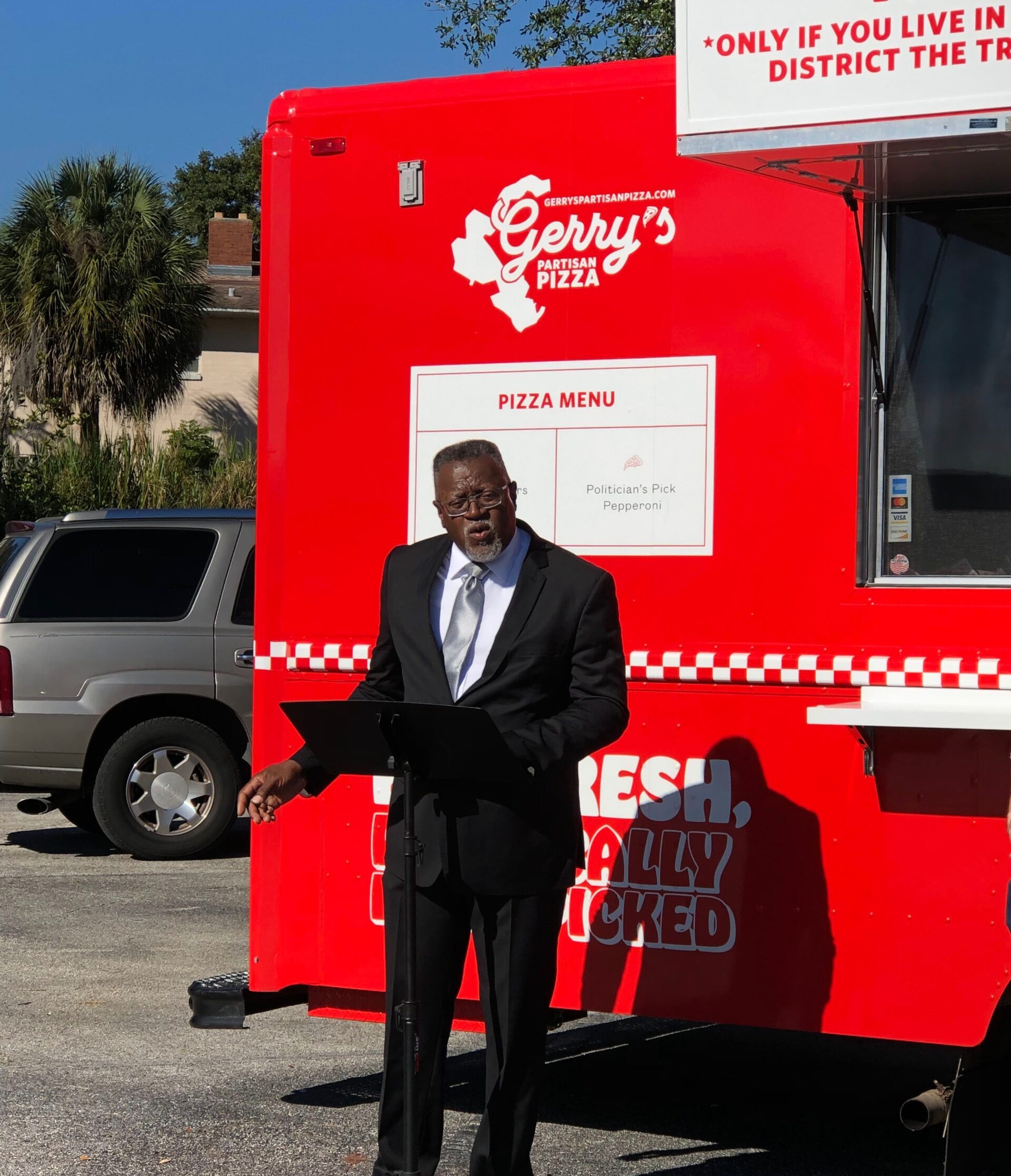 Man in black suit speaks at podium before Gerry's Partisan Pizza food truck with menu outside.