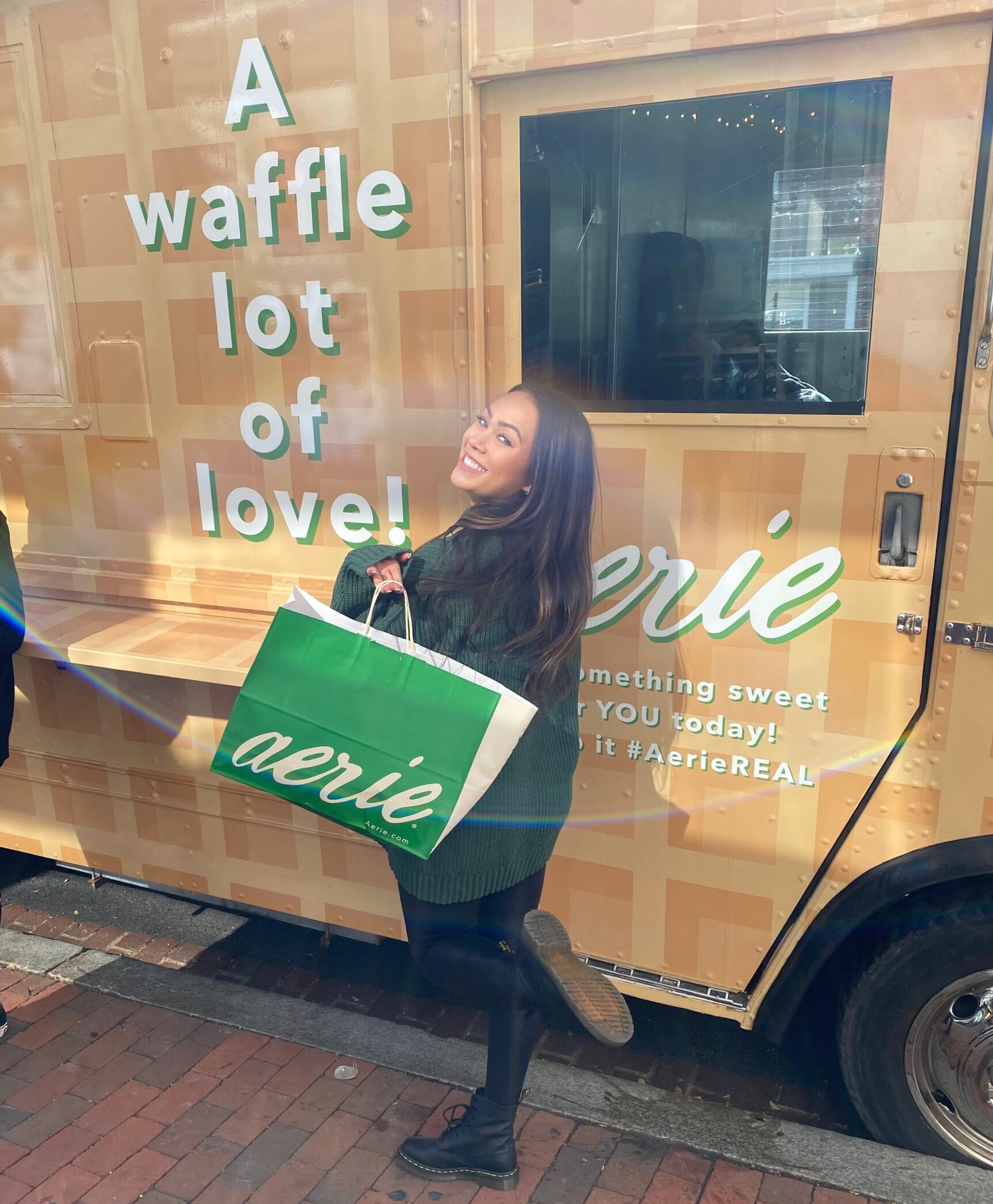 Woman stands by tan waffle truck, holding large green shopping bag on a brick sidewalk.
