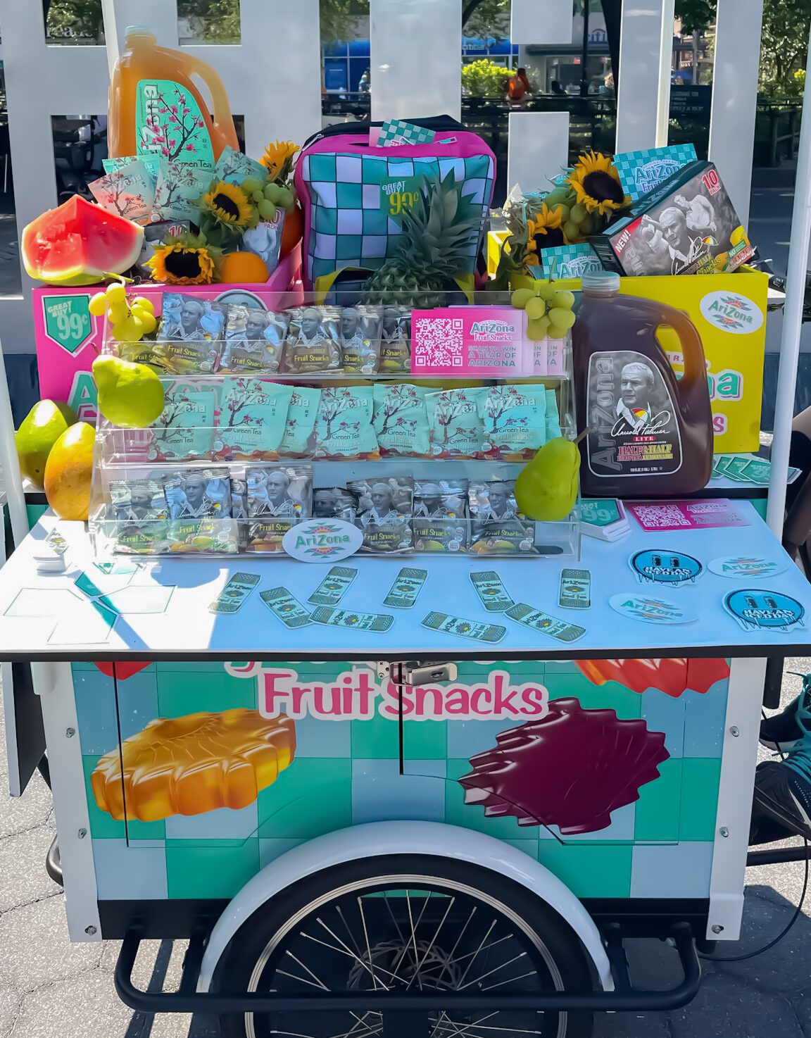 Fruit snack cart with fruit, dollar bills, sunflowers, candies, drinks, and a backpack outdoors.