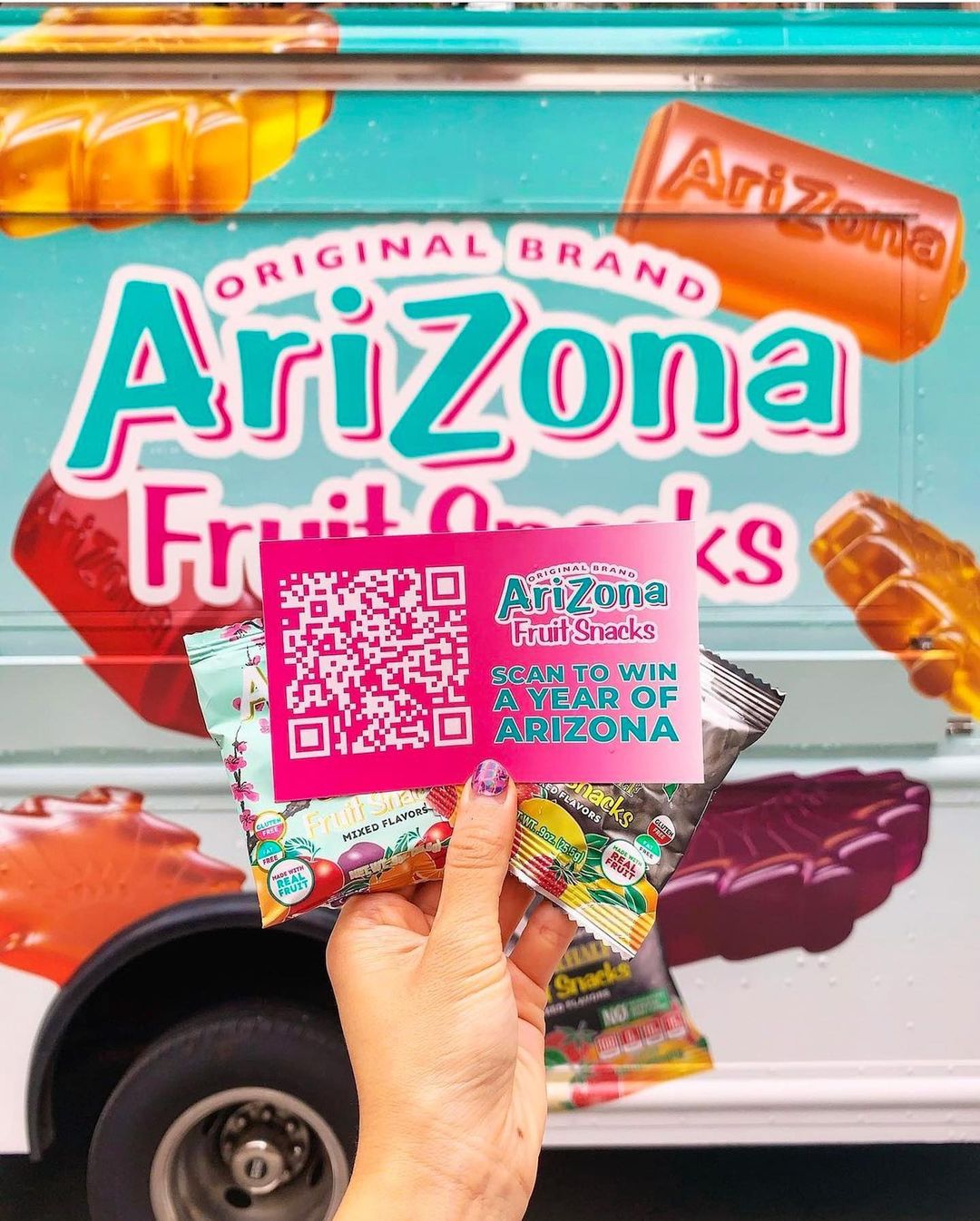 Hand holding two AriZona Fruit Snacks and a QR code card by an AriZona truck at a launch event.