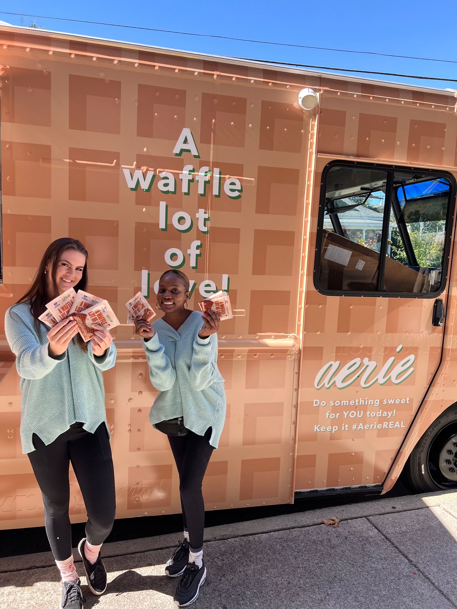 Two women in blue sweaters hold waffle packages in front of a peach waffle truck with "aerie" on it.