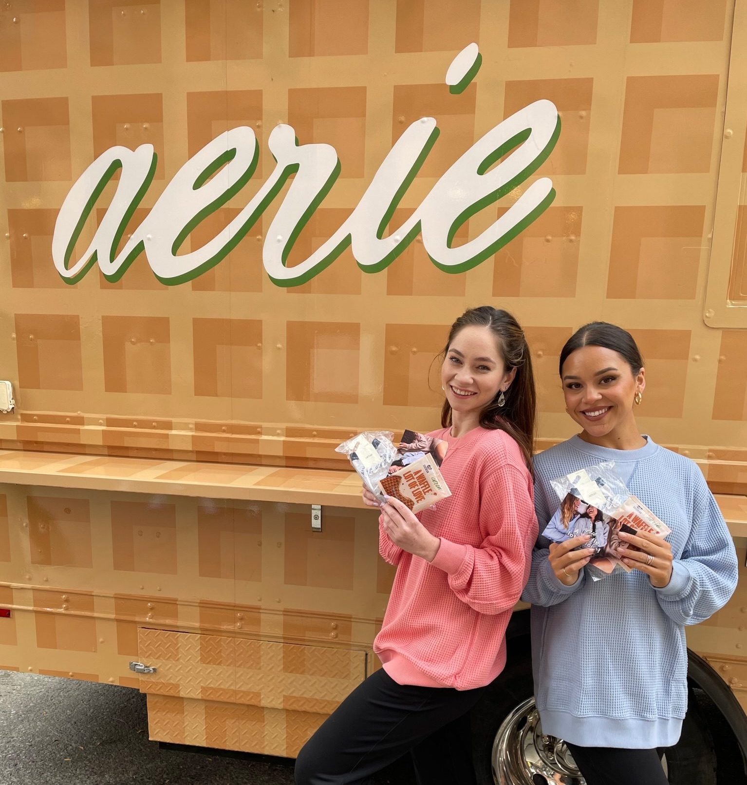 Two women in pink and blue sweaters hold snack packages in front of a tan aerie waffle truck.