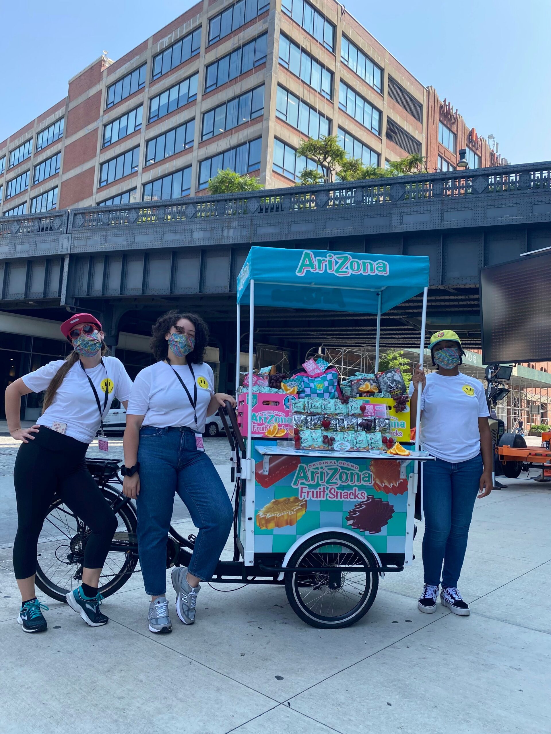 Three people in masks and white shirts stand by an Arizona fruit snacks cart on a city sidewalk.