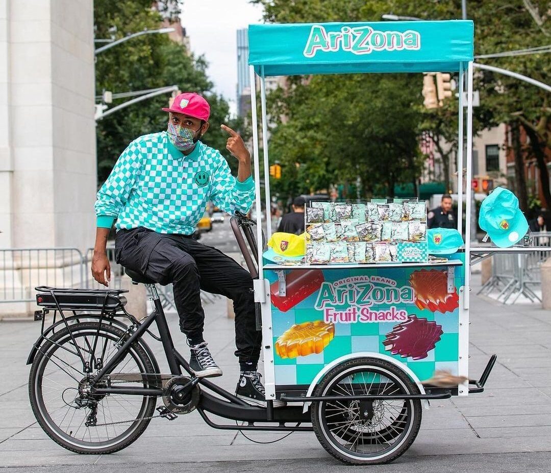 Man in red cap sells AriZona fruit snacks from branded tricycle cart in a city park.