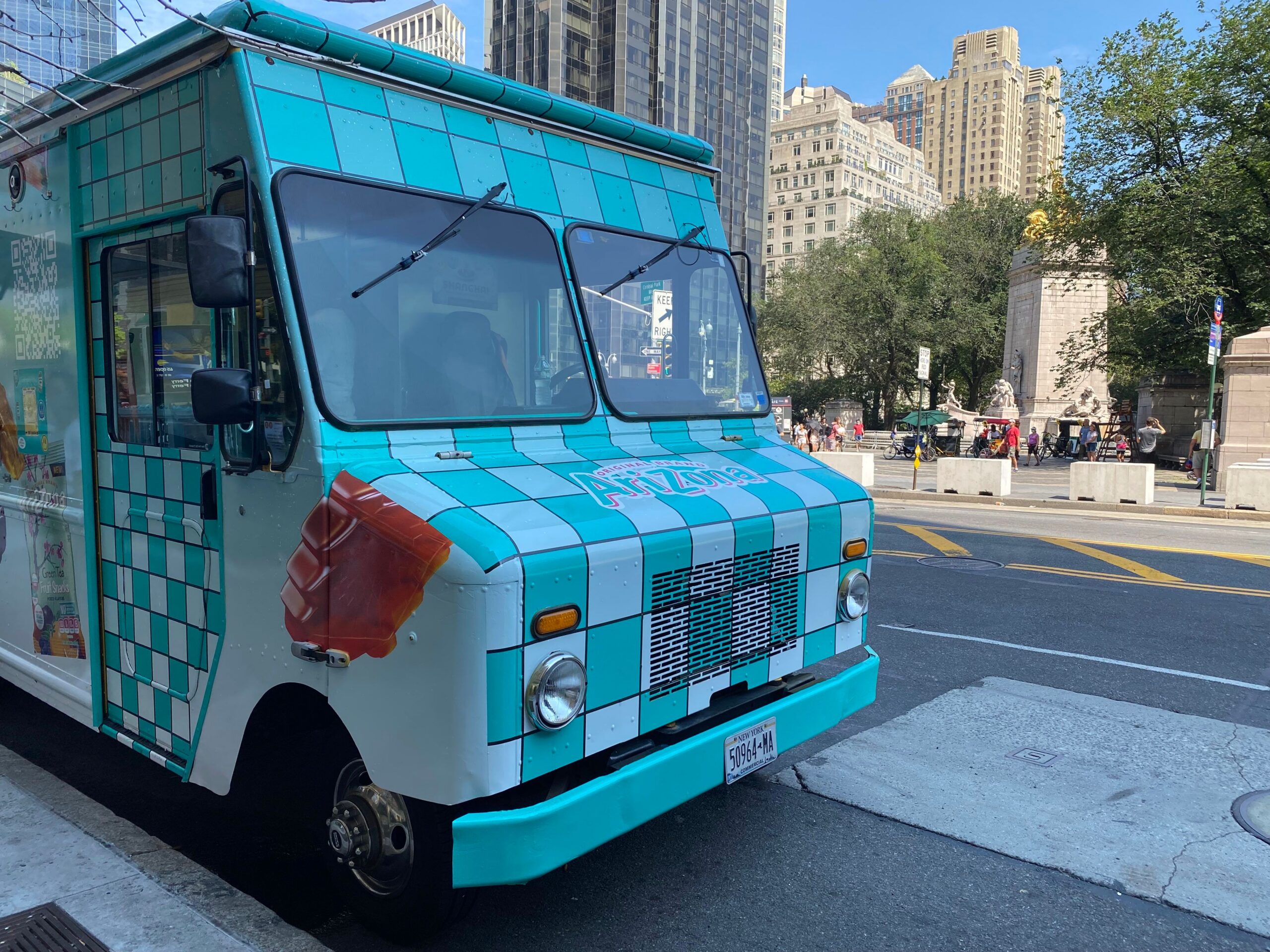 Turquoise and white ice cream truck with cone graphic parked by tall buildings and a park.