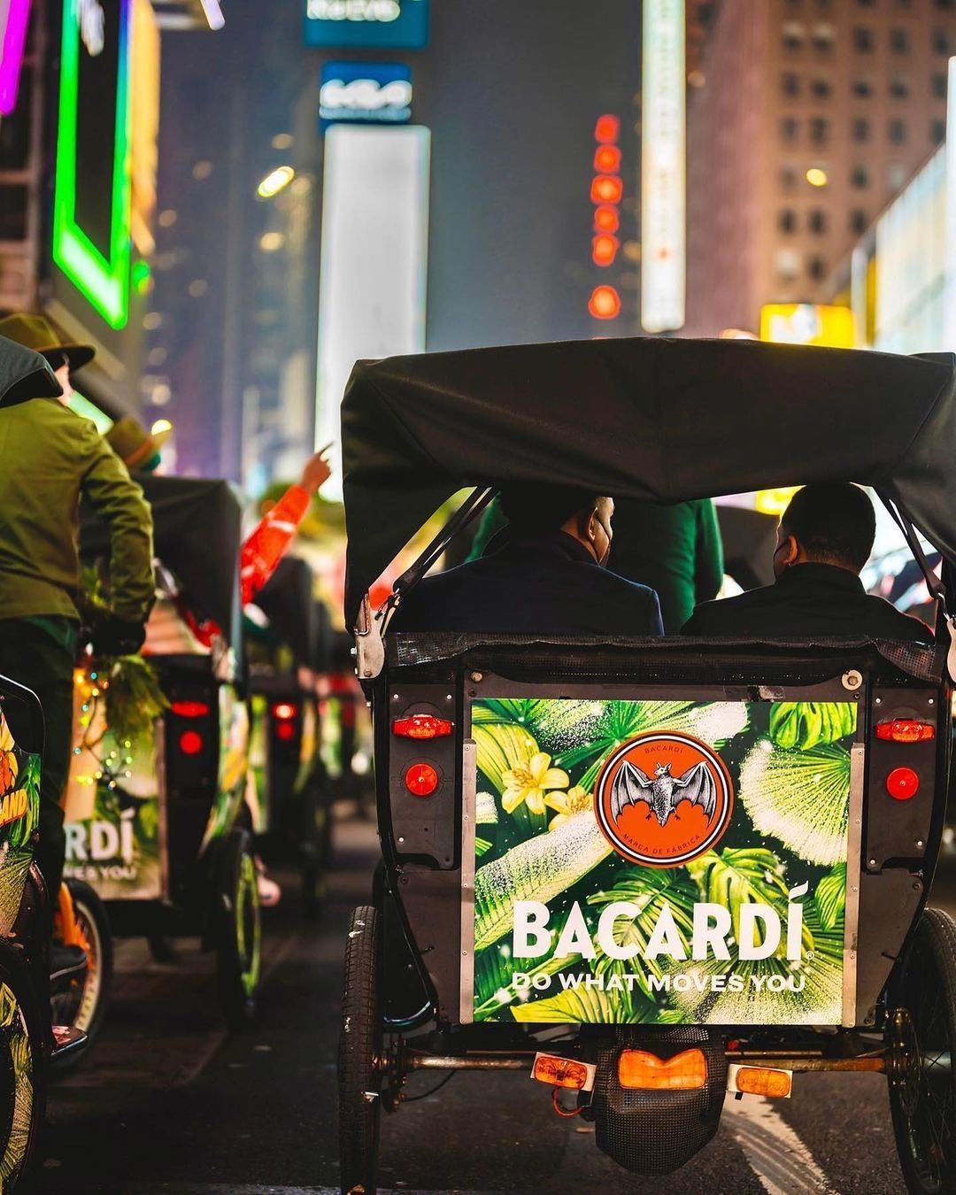 Pedicabs with a Bacardi ad ride through city streets lit by neon and holiday decorations.