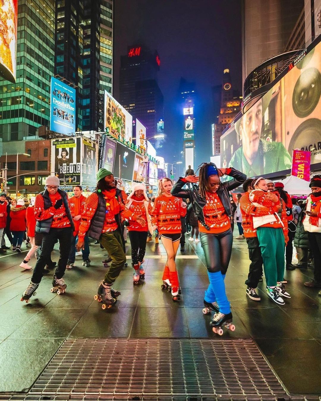 People in matching red sweaters roller skate at night in Times Square amid bright lights and crowds.