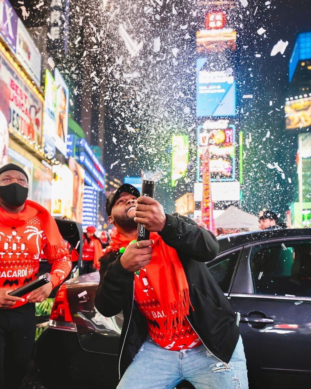 Man in red scarf pops confetti cannon at a holiday pop-up in Times Square at night.