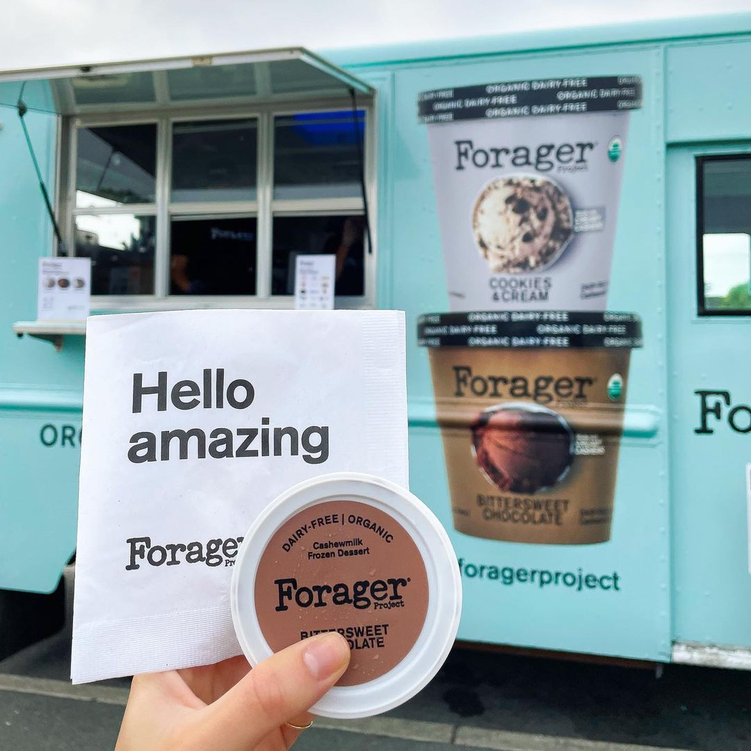 Hand holding a “Hello amazing” napkin and Forager chocolate dessert by a blue food truck.