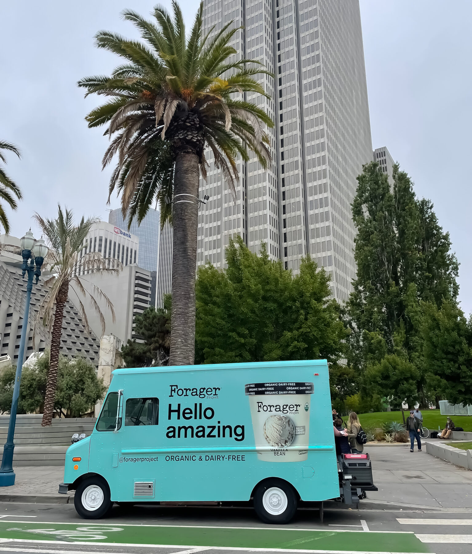 Turquoise Forager food truck offers organic, dairy-free samples on a city street with palm trees.