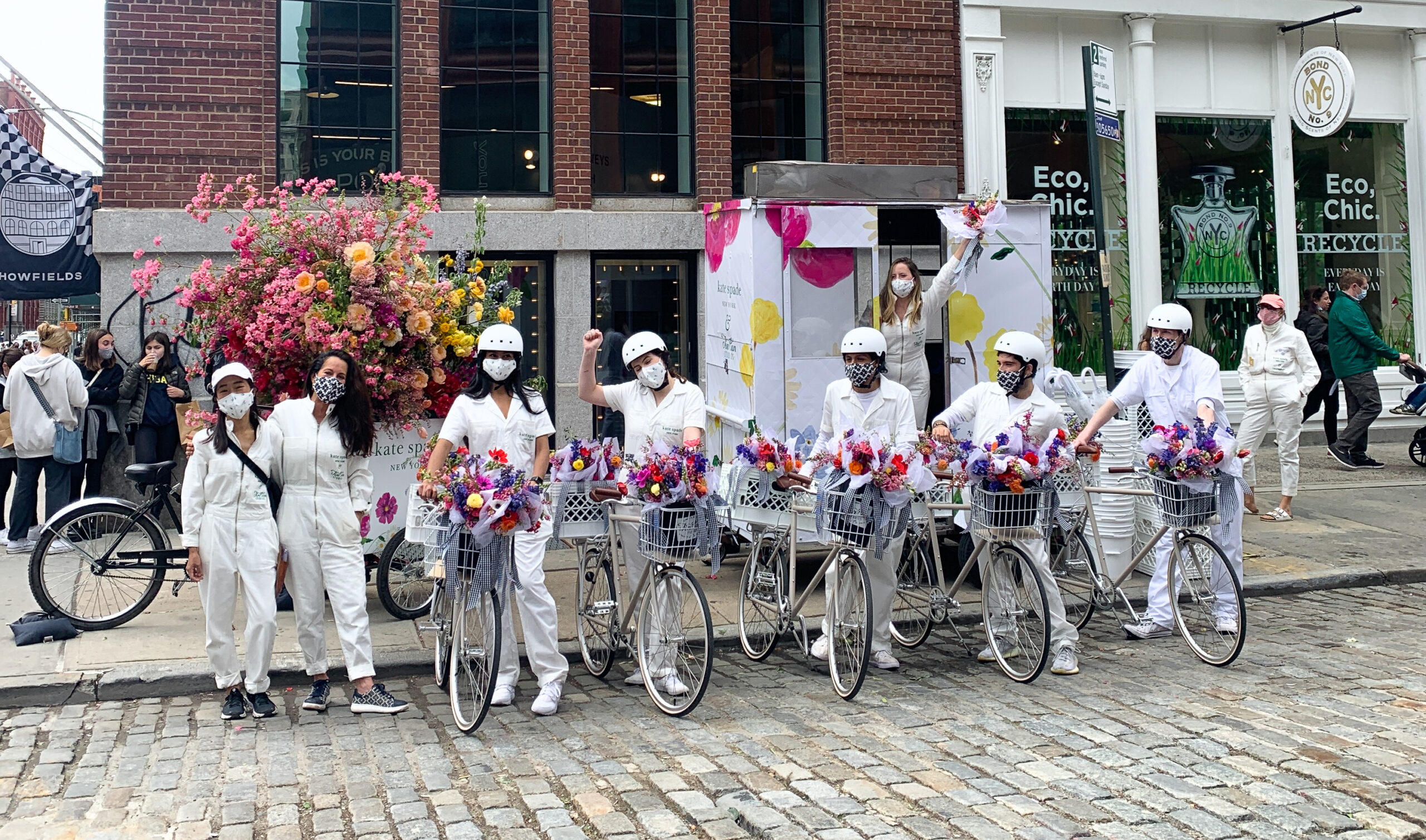 People in white with helmets and masks stand by flower-decorated bikes on a cobblestone street.