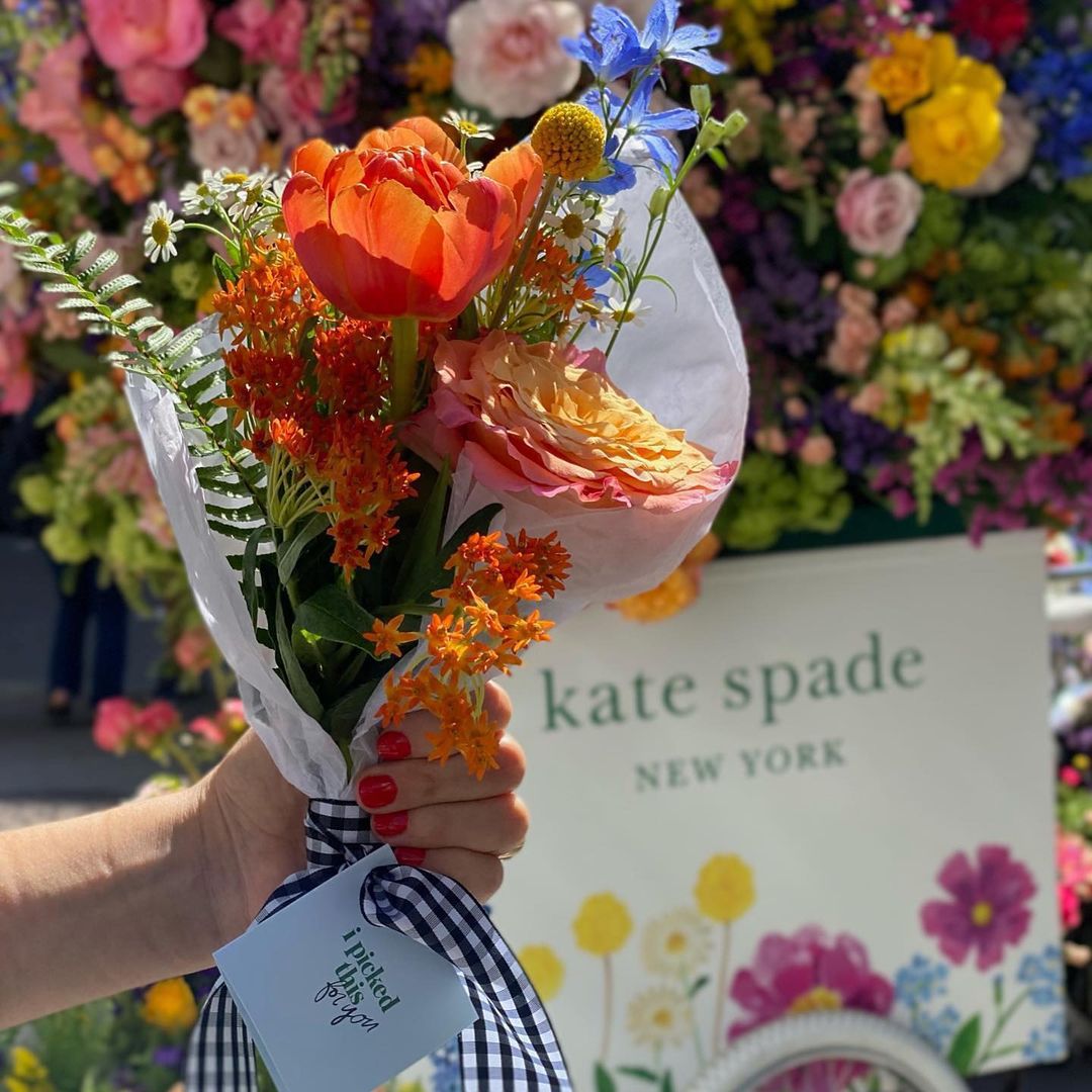 Hand with red nail polish holds a flower bouquet in front of kate spade new york holiday sign.