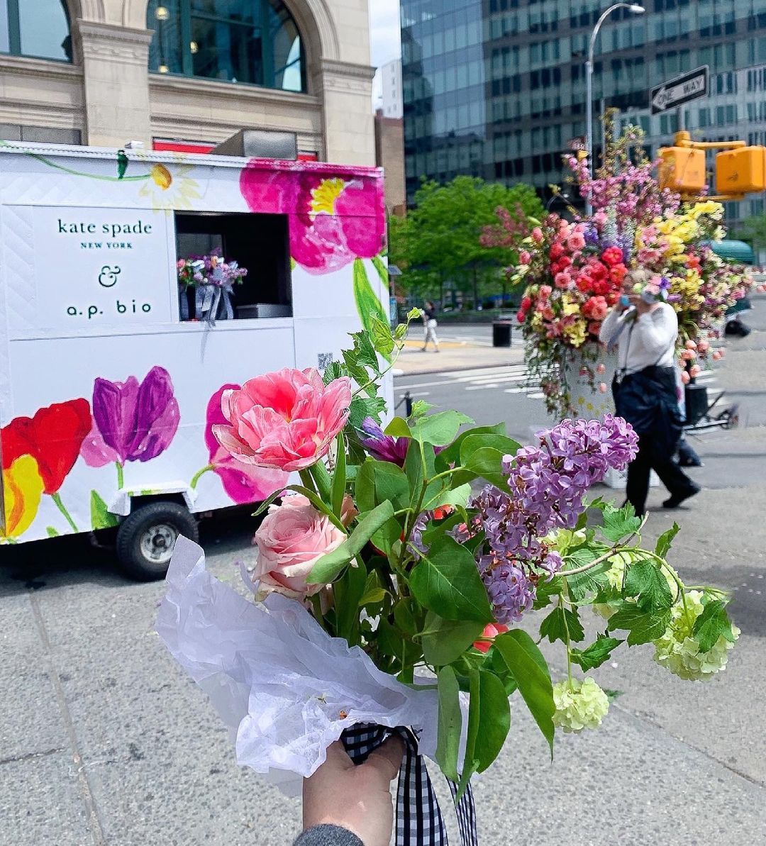 A hand holds a bouquet by a floral food truck; nearby, a man carries flowers at a city intersection.