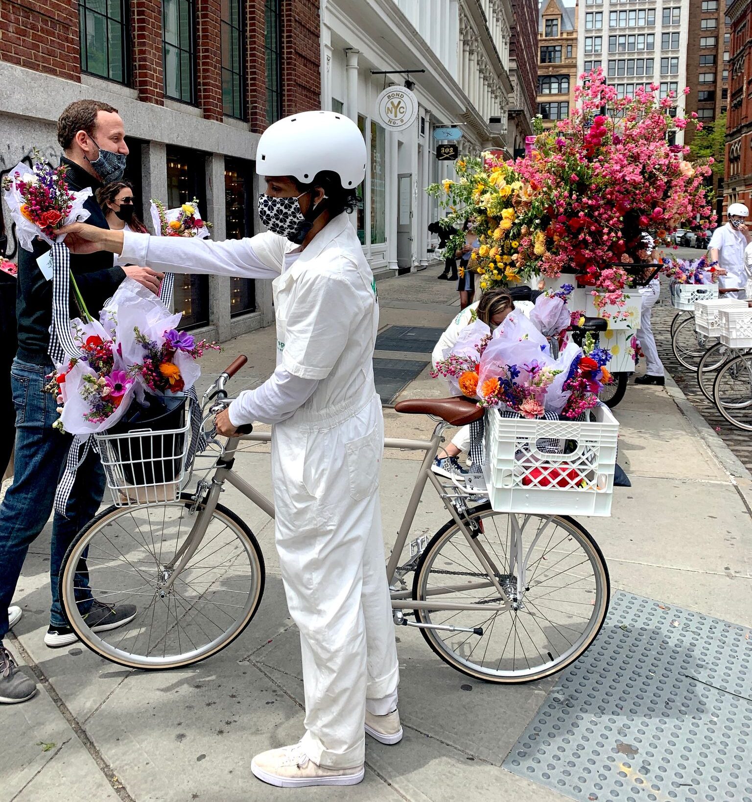 People in white coveralls give flower bouquets from bikes to passersby on a city sidewalk.