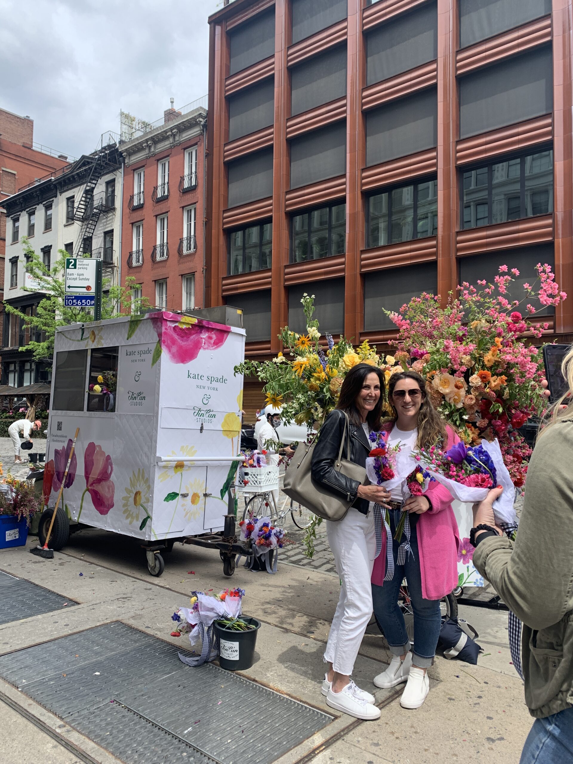 Two women hold floral bouquets by a kate spade flower cart on a city sidewalk with tall buildings.