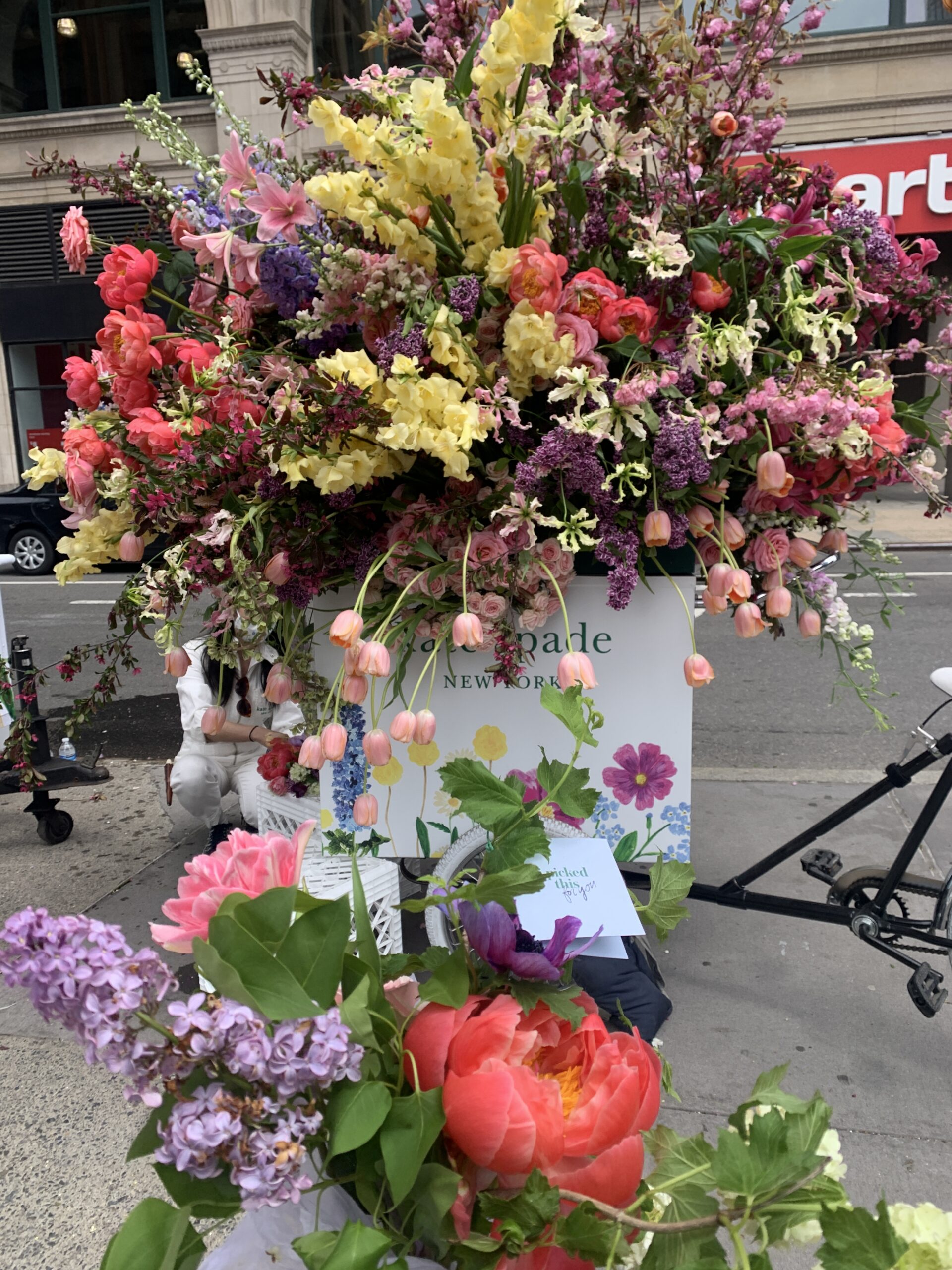 Pink, purple, yellow, and red flowers overflow a Kate Spade New York display on a city sidewalk.