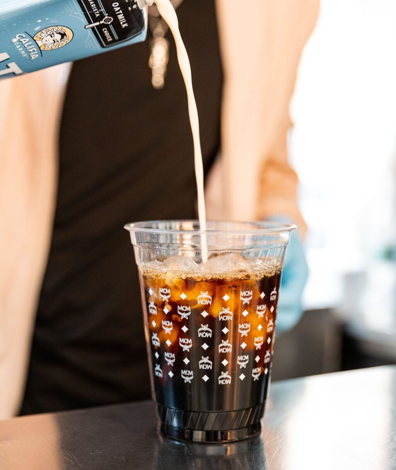 Person in blue glove pours milk into iced coffee cup with logo at MCM pop-up café.