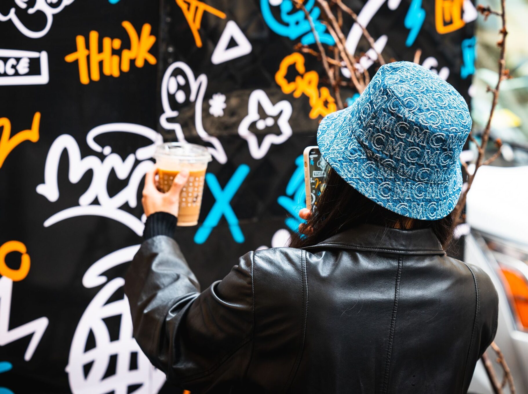 Person in blue bucket hat holds iced coffee and photographs colorful graffiti wall with phone.