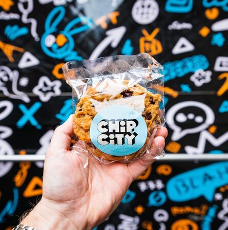 Hand holds a clear bag of Chip City chocolate chip cookies in front of a colorful doodle wall.