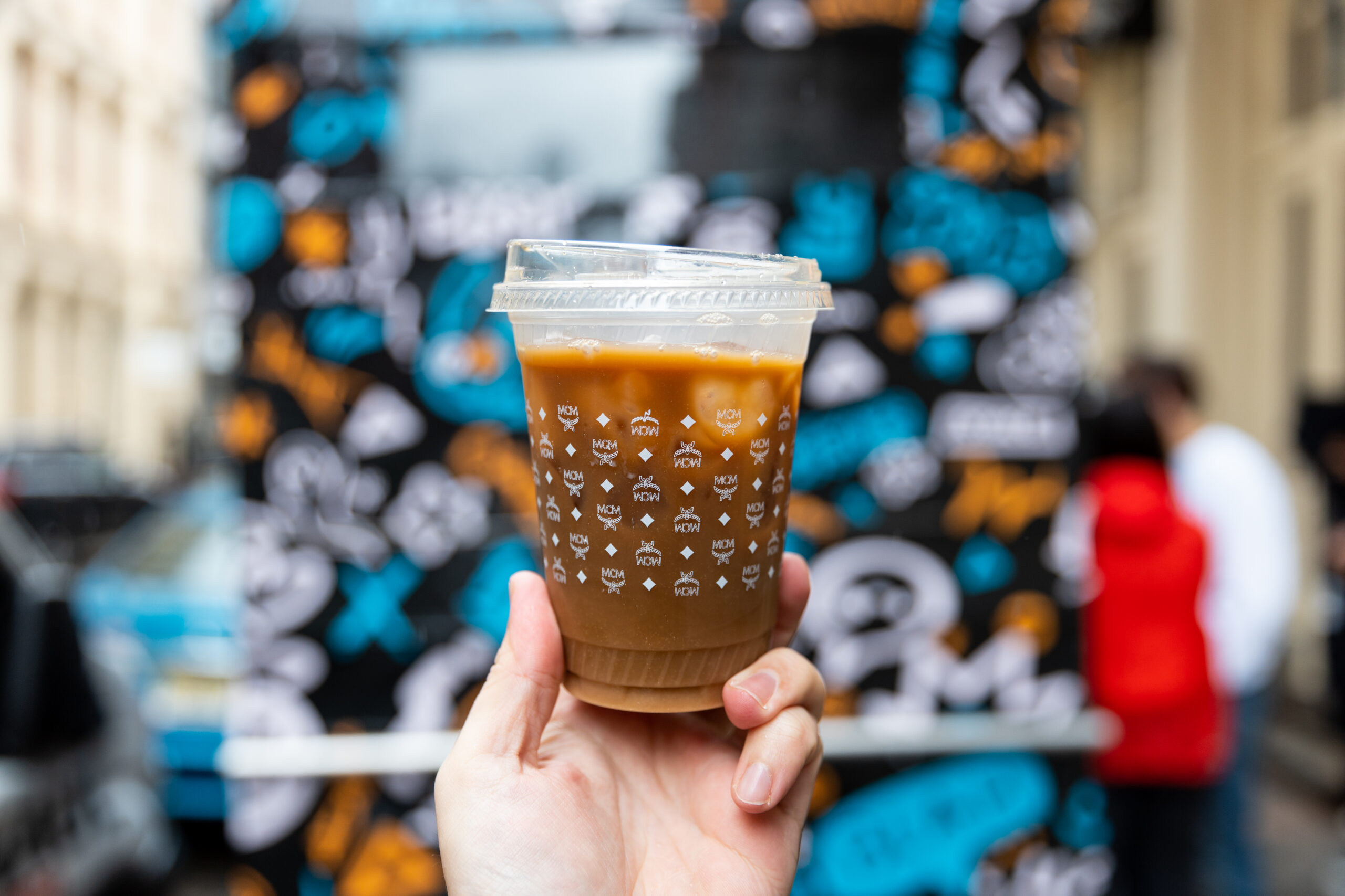 Hand holding iced coffee in clear cup with white designs; blurred graffiti and people behind.