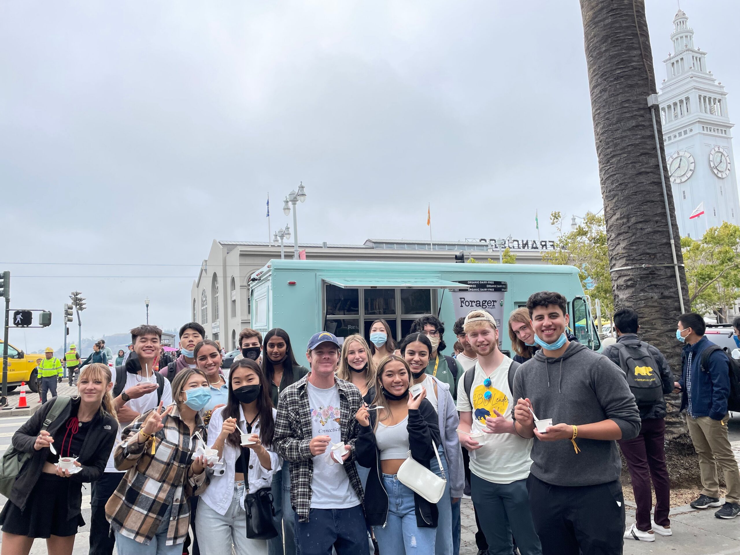 Young adults with ice cream stand by a blue food truck, some masked; clock tower in back.