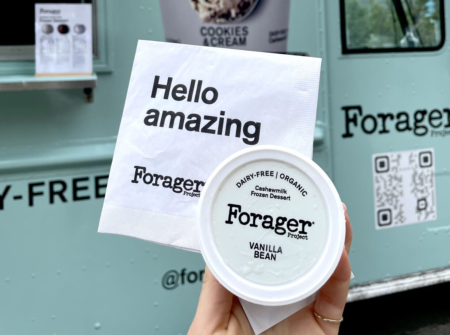 Hand holding Forager Project vanilla bean cashewmilk dessert and napkin near mint green food truck.