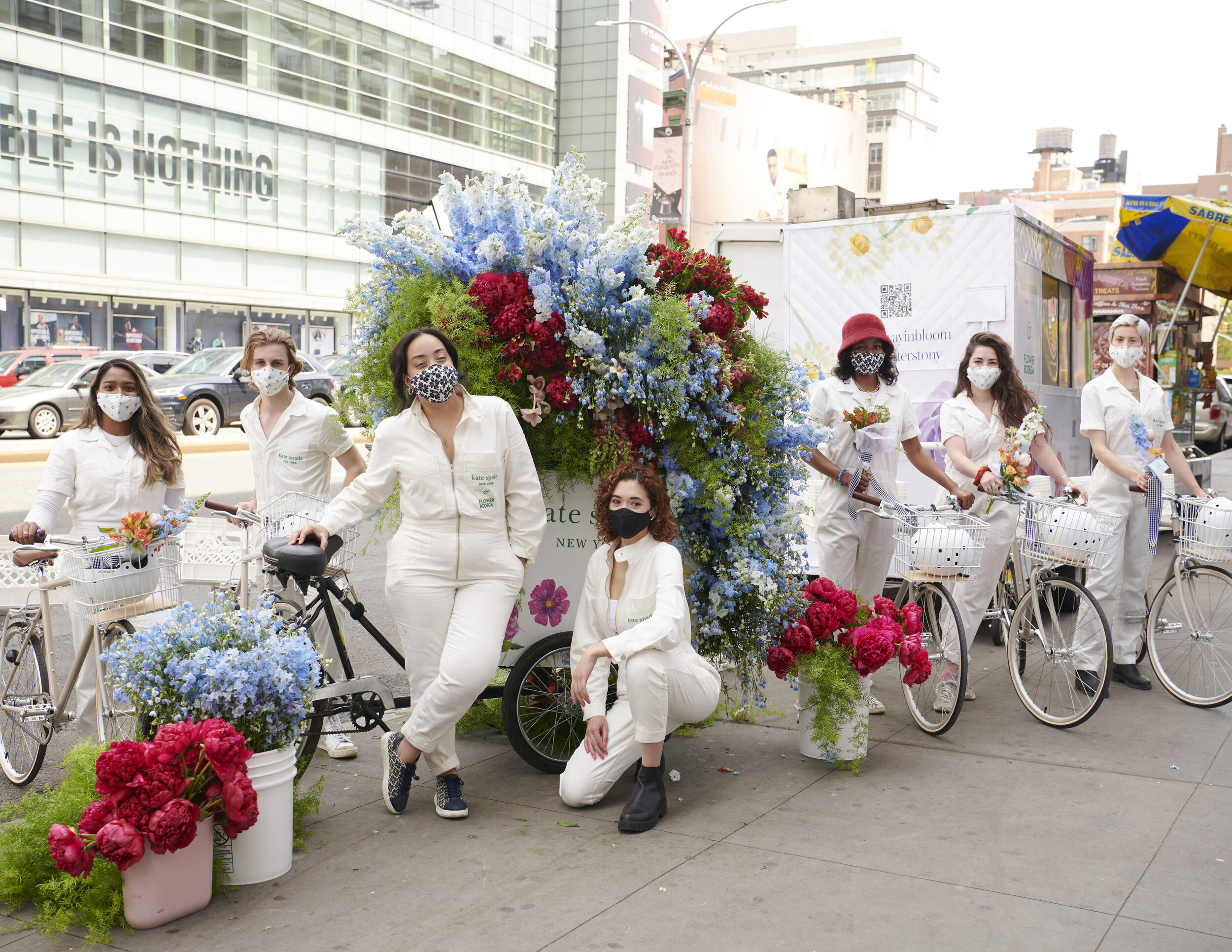 Seven people in white and masks pose with white bikes and bright flowers on a city sidewalk.