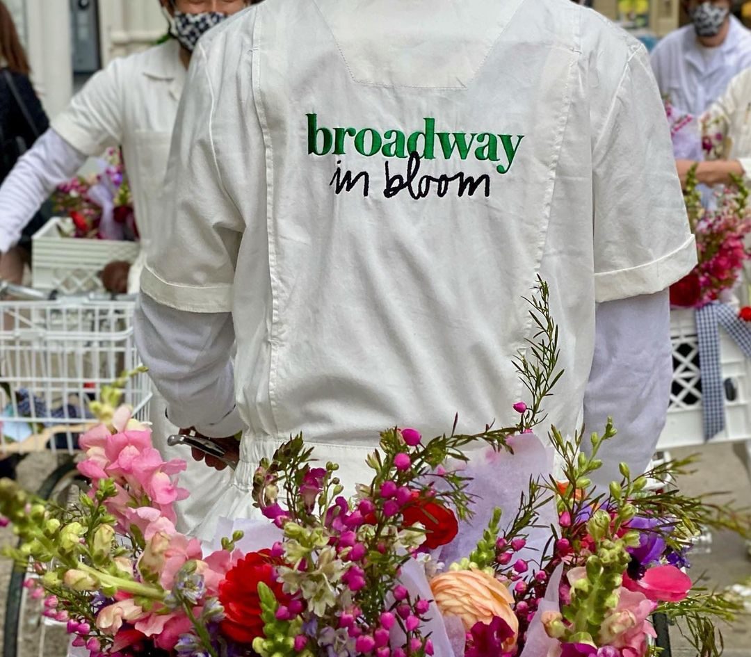 Person in white with "broadway in bloom" on back stands by flower baskets at holiday pop-up.