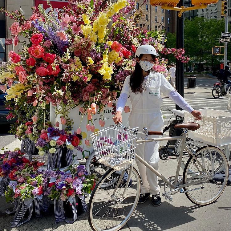 Woman in white overalls and helmet stands by a bike next to a pop-up floral display on sidewalk.