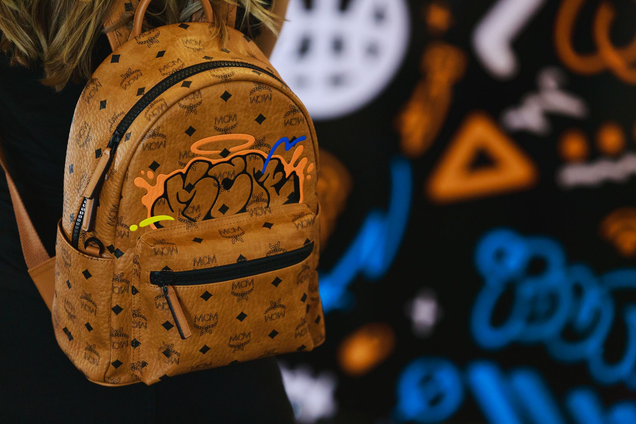 Tan leather MCM backpack with black monogram and colorful graffiti, worn against a blurred mural.