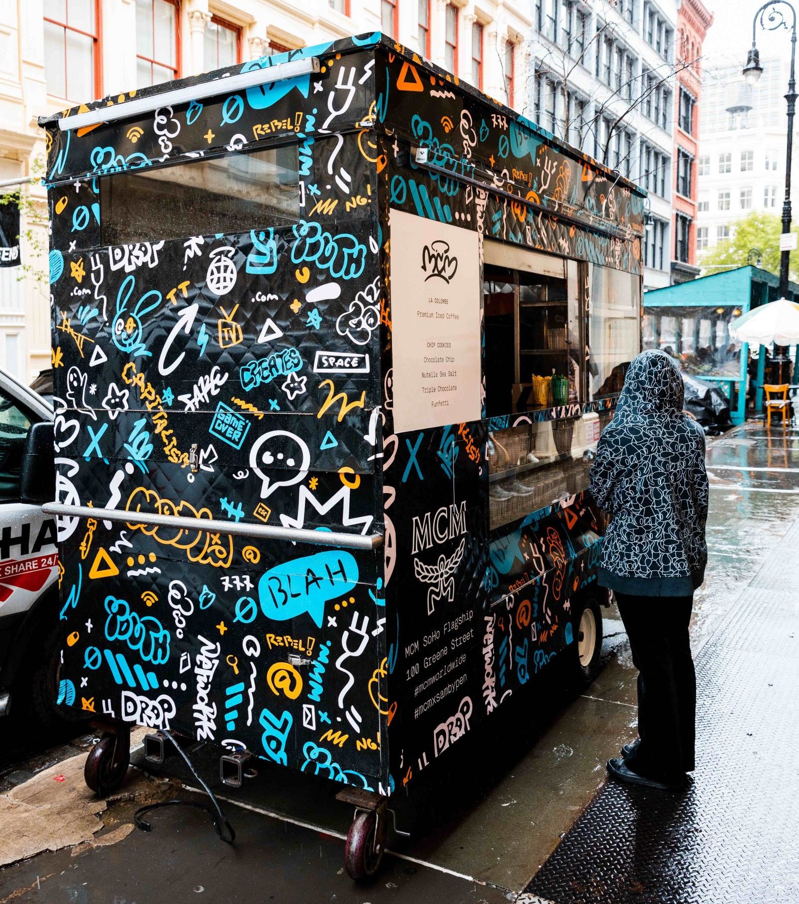 Person in a hooded jacket at a graffiti-covered food cart on a rainy city street.