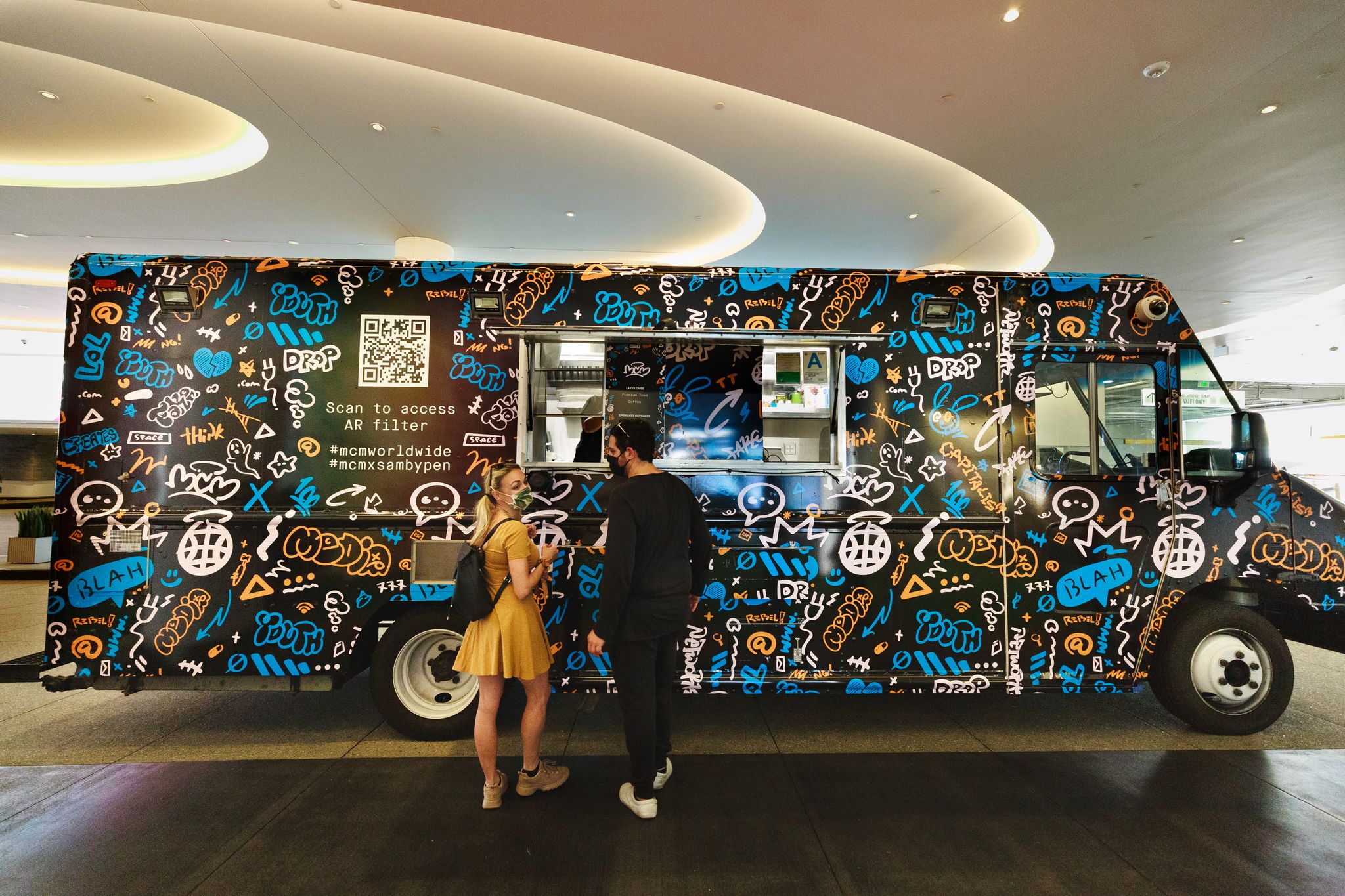 MCM café food truck with graffiti designs and a QR code serves two people indoors.
