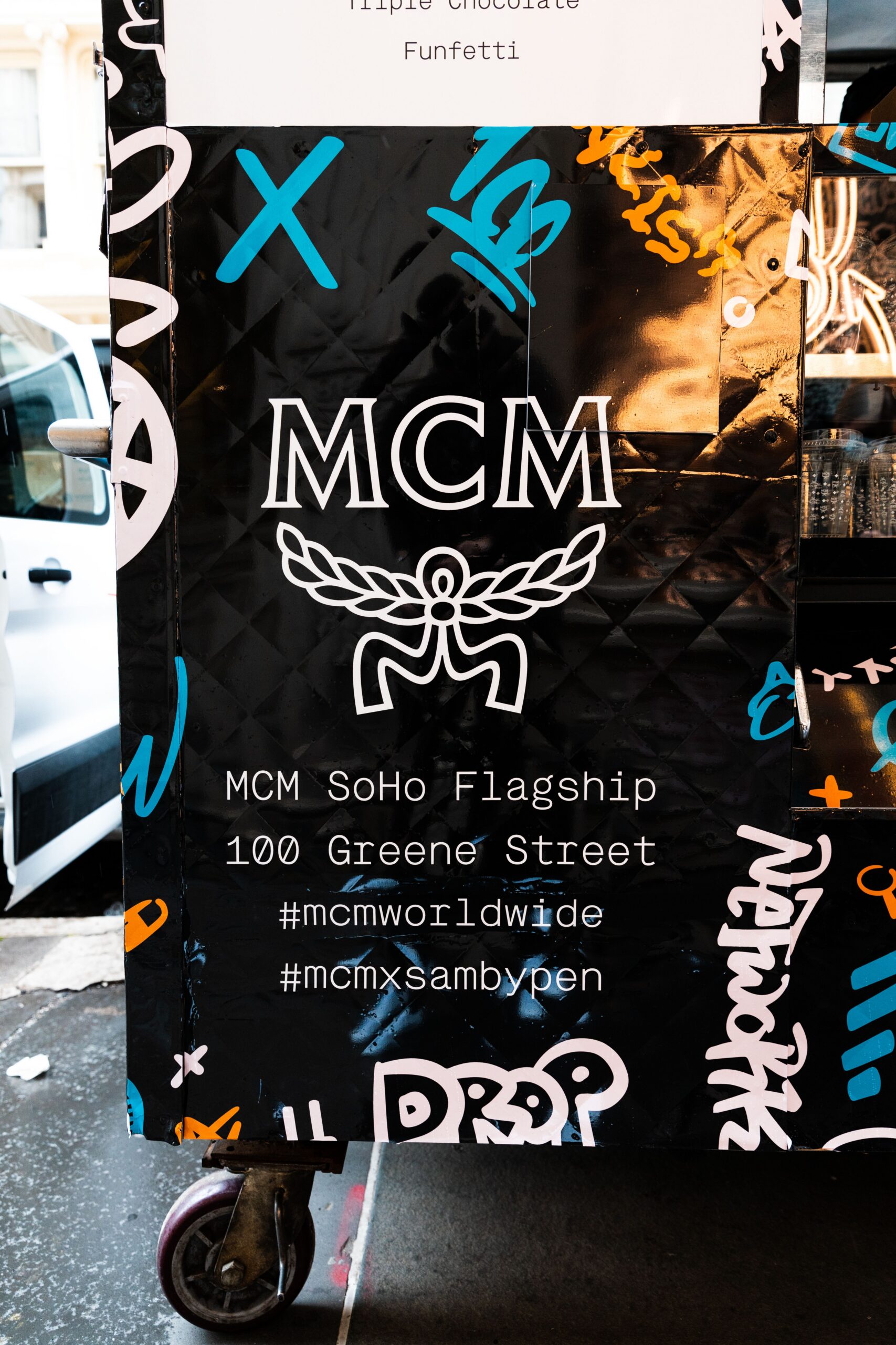 Black quilted panel with MCM logo, stylized wings, white graffiti designs, and event text.