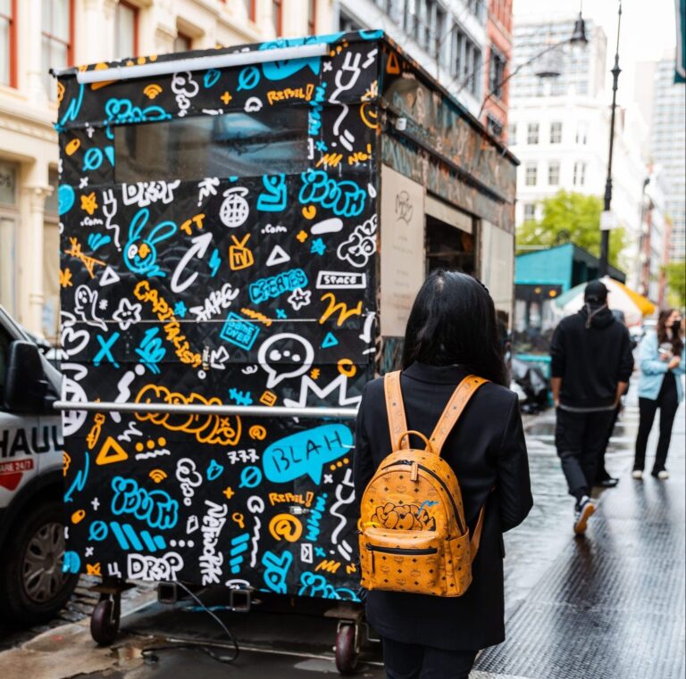 Person with long dark hair and orange backpack walks toward a colorful graffiti MCM food truck.