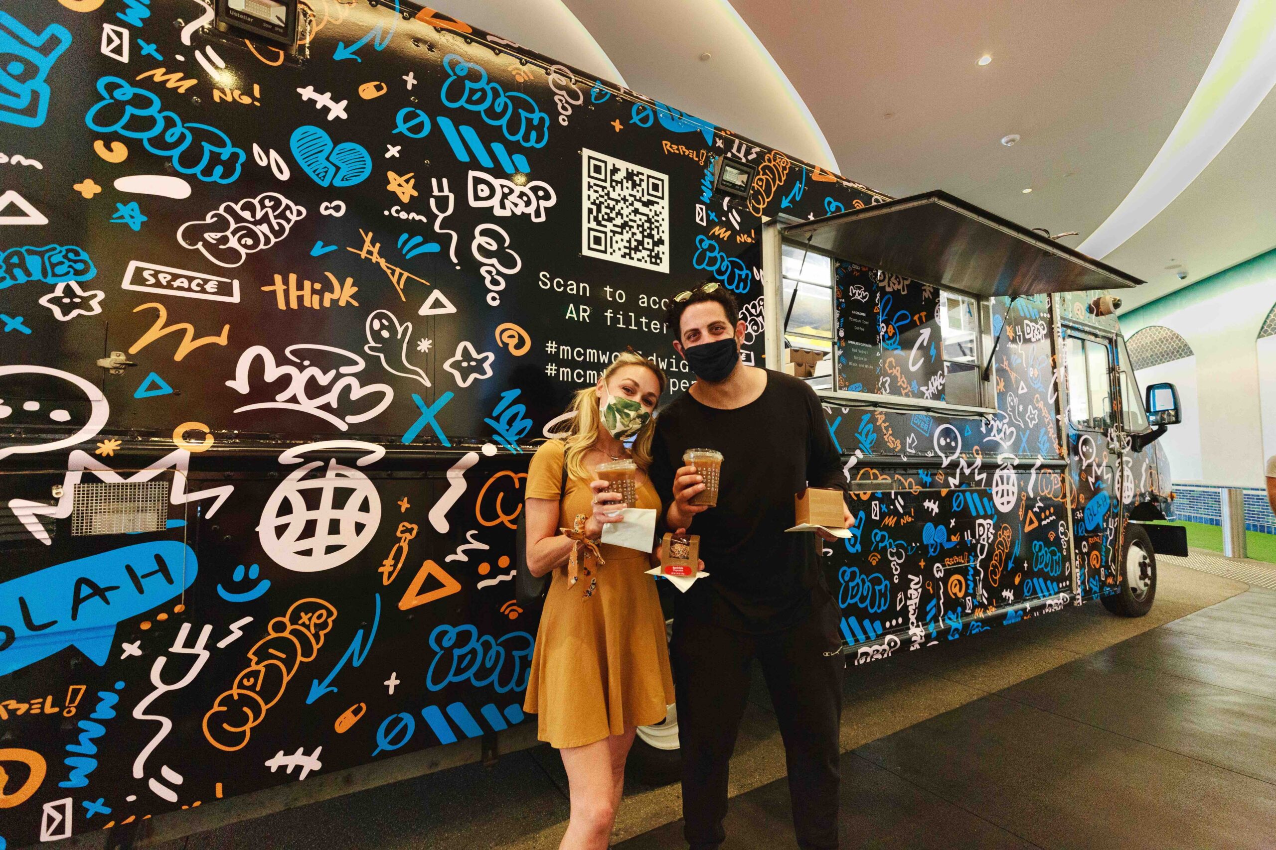 Two masked people stand by a colorful MCM food truck with graffiti, holding drinks and food.