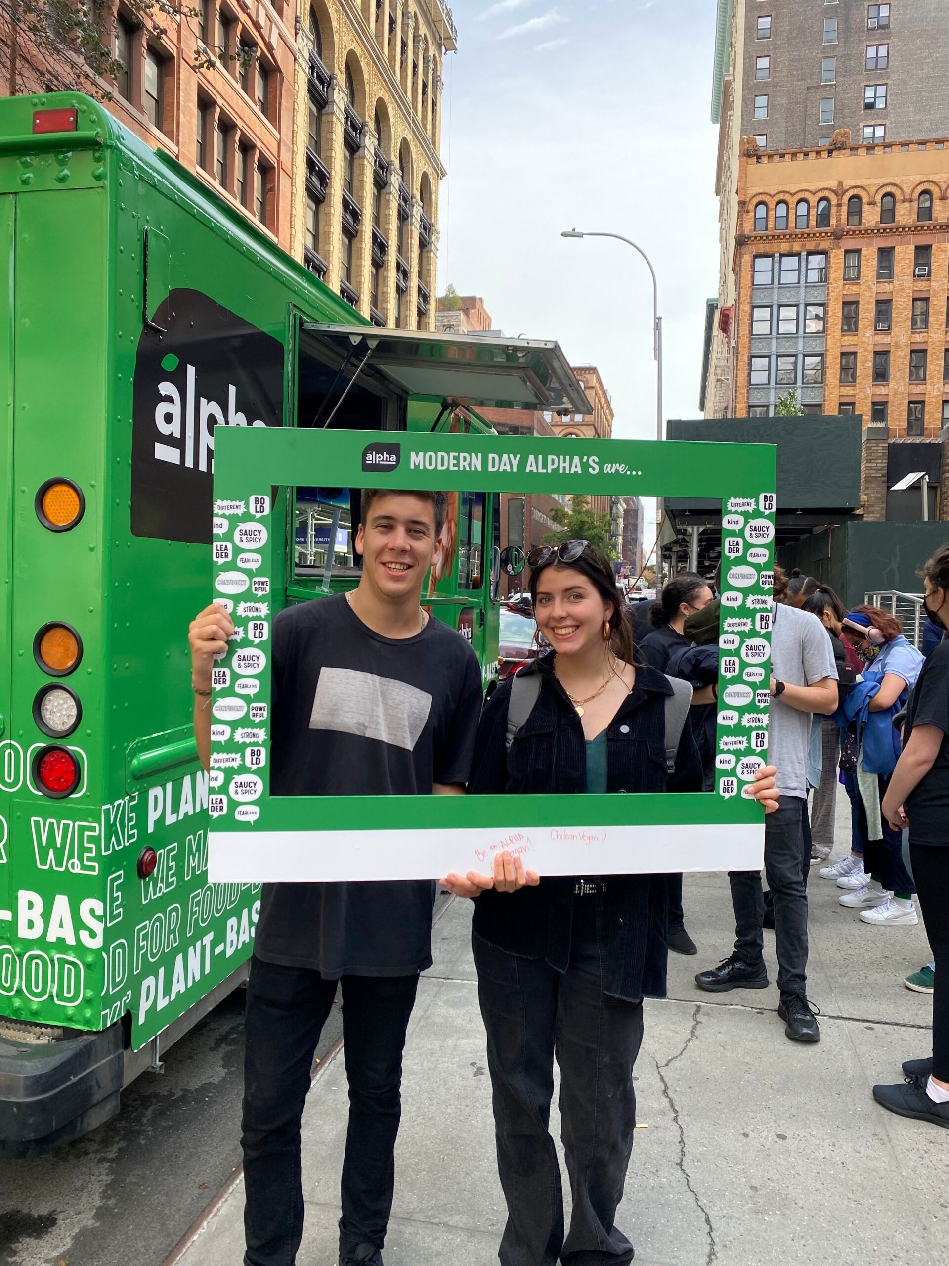 Two people smile on a city sidewalk, holding a "Modern Day Alpha's" frame near a green food truck.