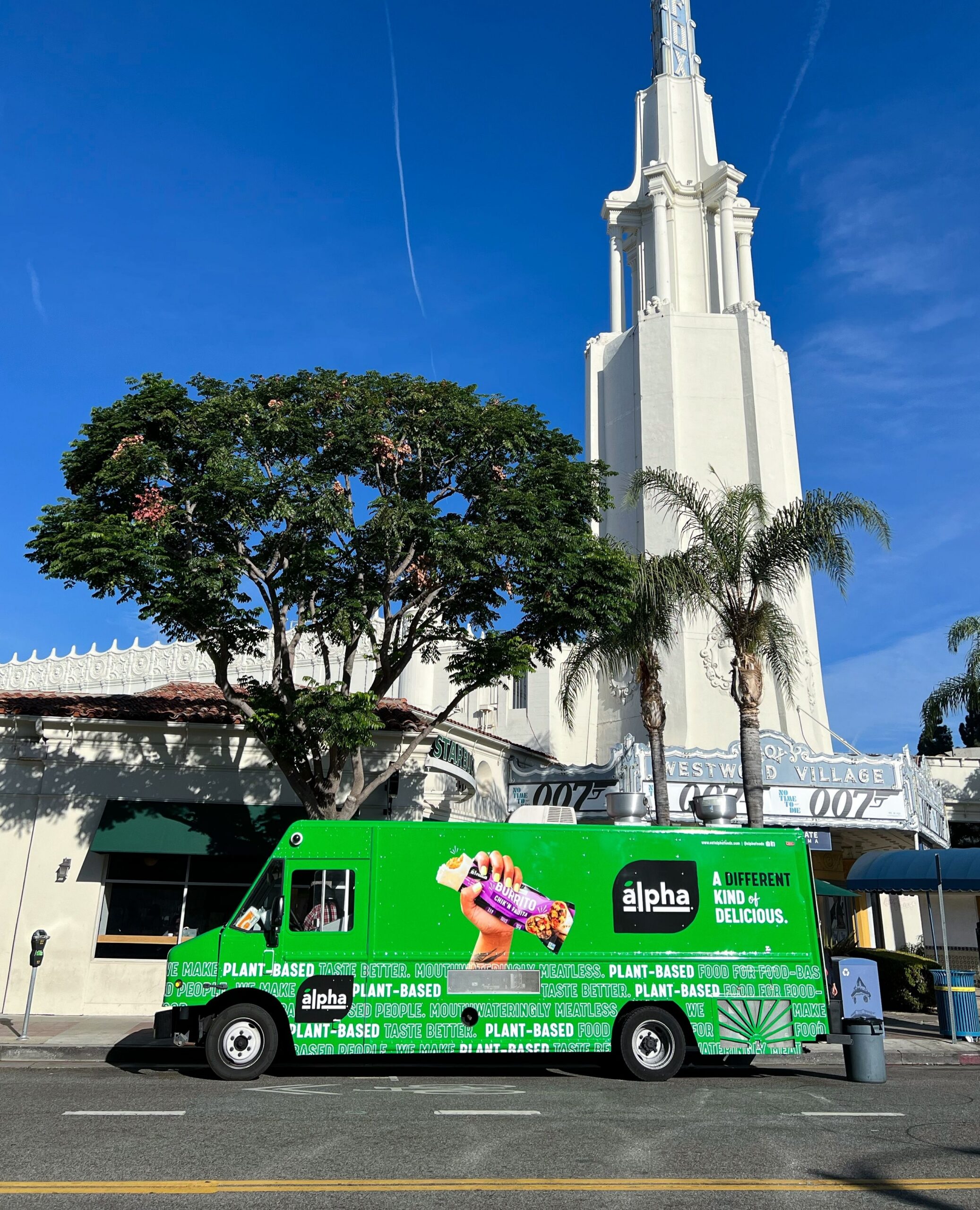 Green Alpha Foods food truck with plant-based signs parked by a white building and palm trees.