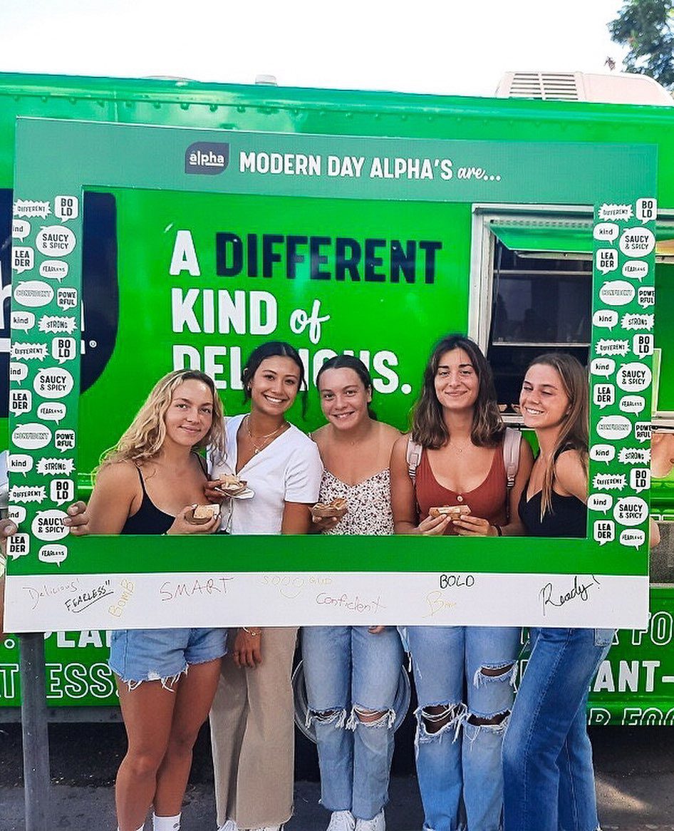 Five young women smile holding food in front of a green food truck and large photo frame.