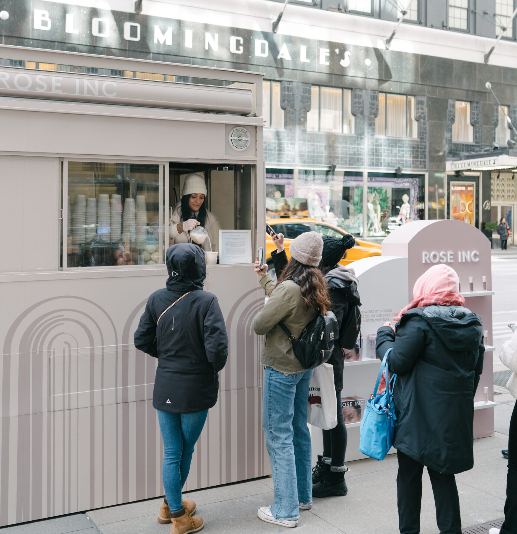 Four people line up at a Rose Inc. kiosk on a city sidewalk as some take photos with phones.