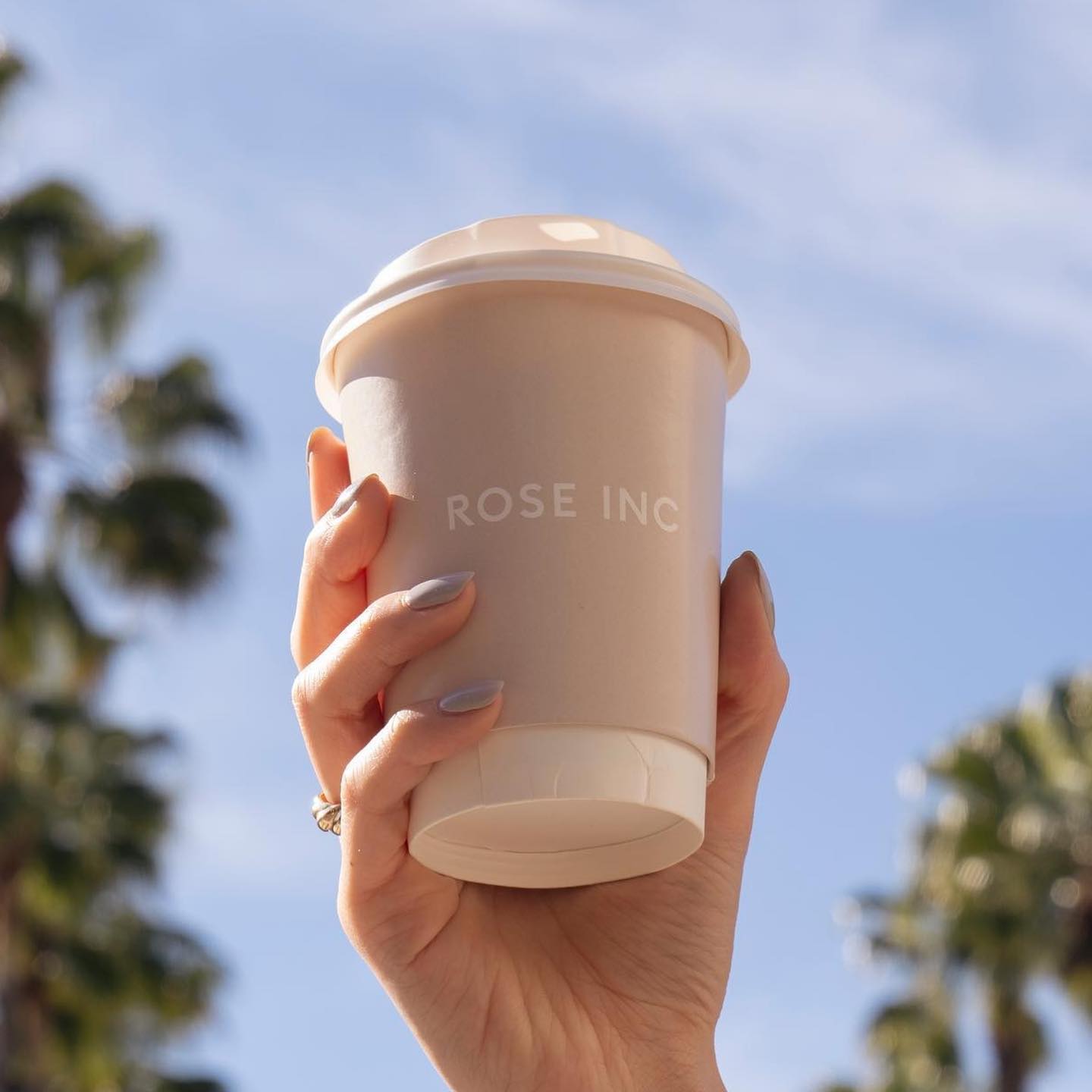 Hand with light purple nails holds a ROSE INC coffee cup outdoors with blue sky and palm trees.