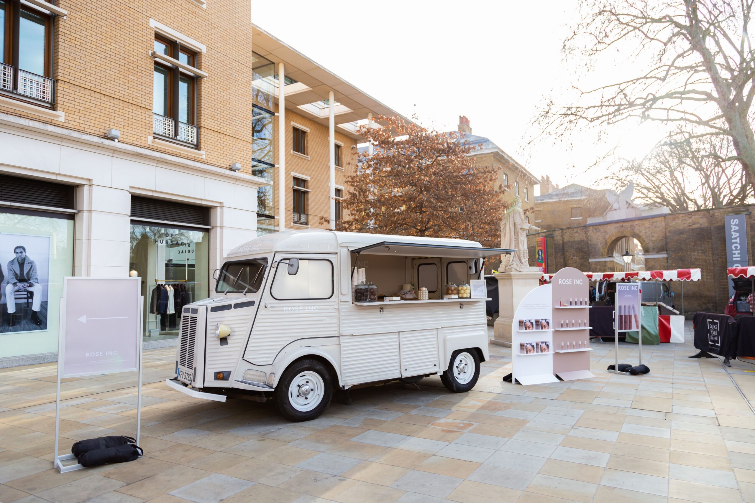 White food truck on a plaza with signs and displays, trees, and modern buildings in the background.