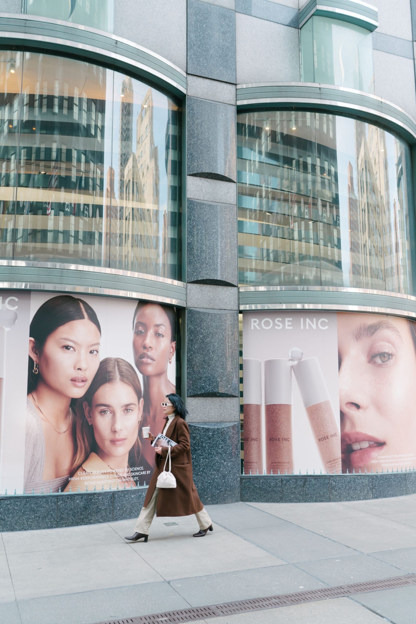 Person in brown coat walks past city building with beauty ads and a pop-up for cosmetics.