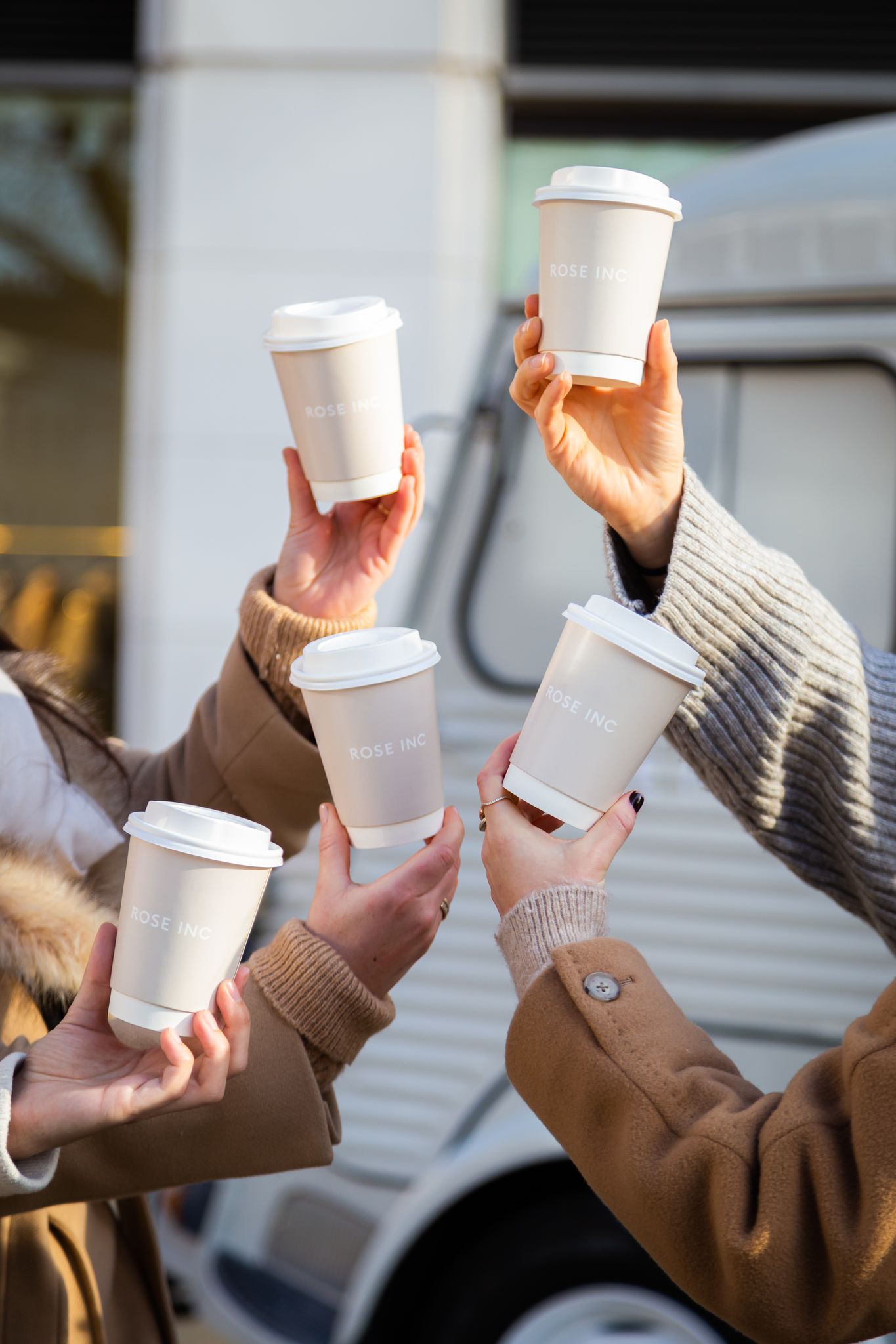 Five hands raise ROSE INC coffee cups together outdoors near a van. All wear coats.