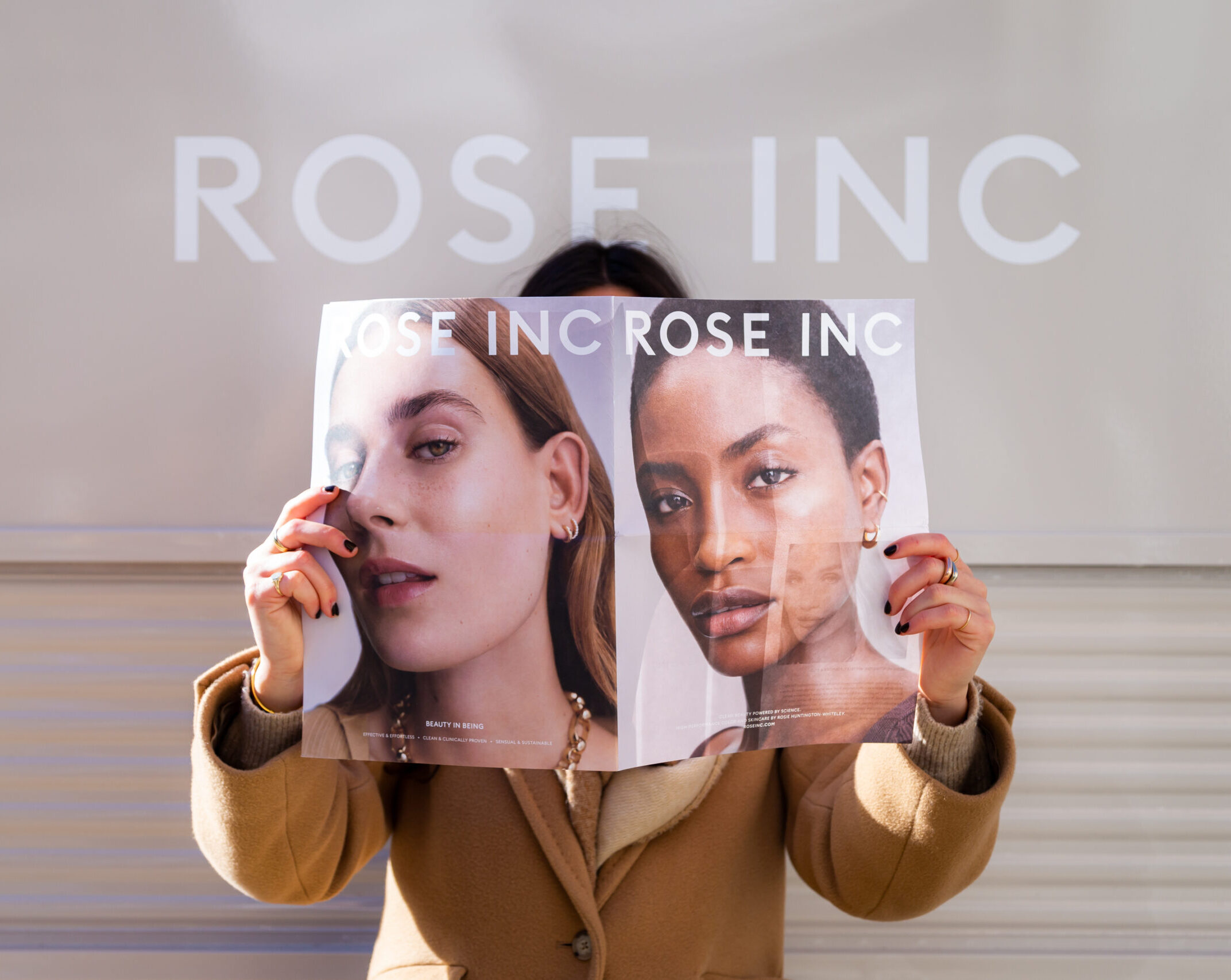 Person in tan coat holds up magazine with two women’s faces, covering their own, by a ROSE INC sign.