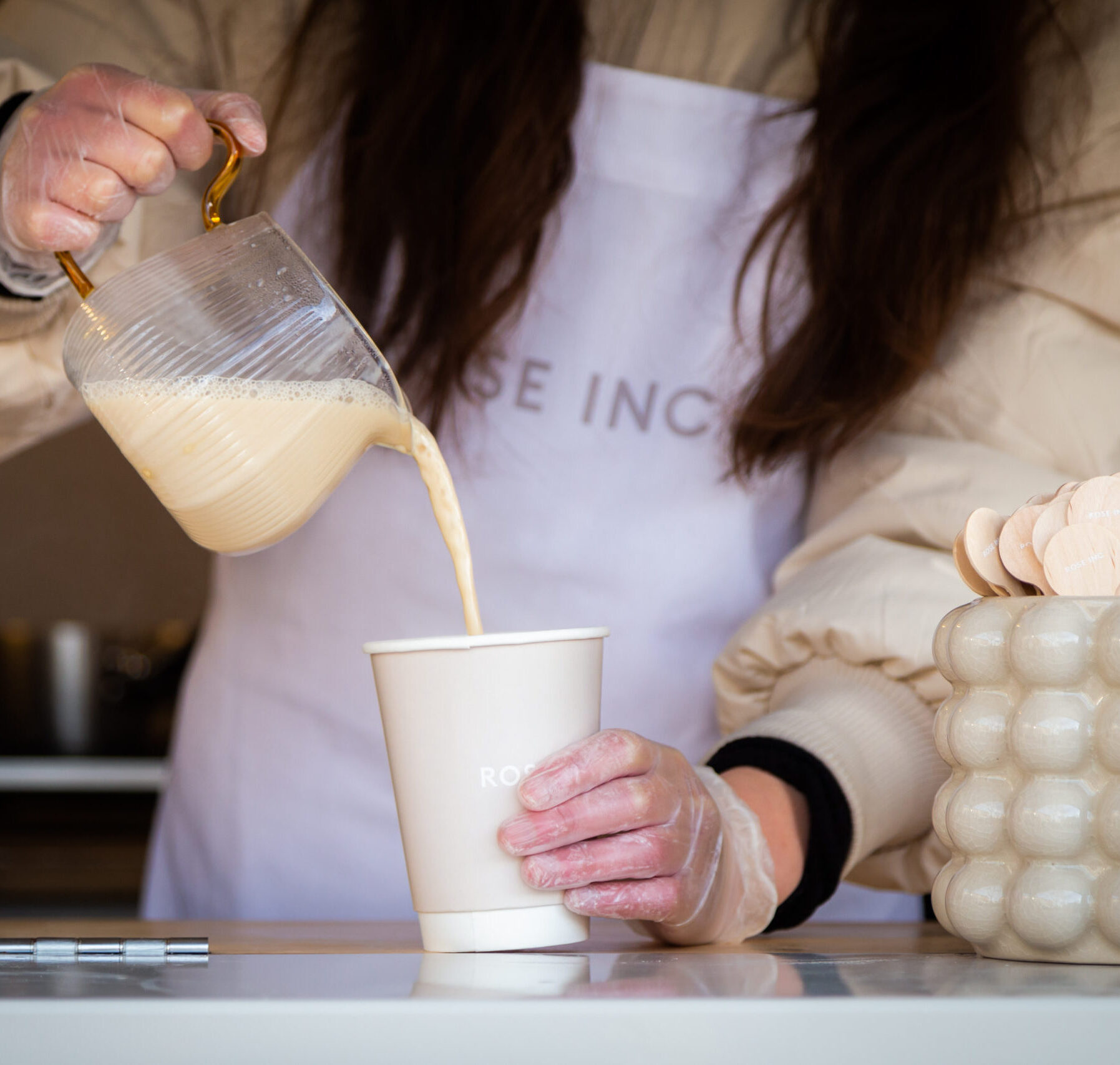 A gloved person pours a beverage from a glass jug into a paper cup beside stirrers and a container.