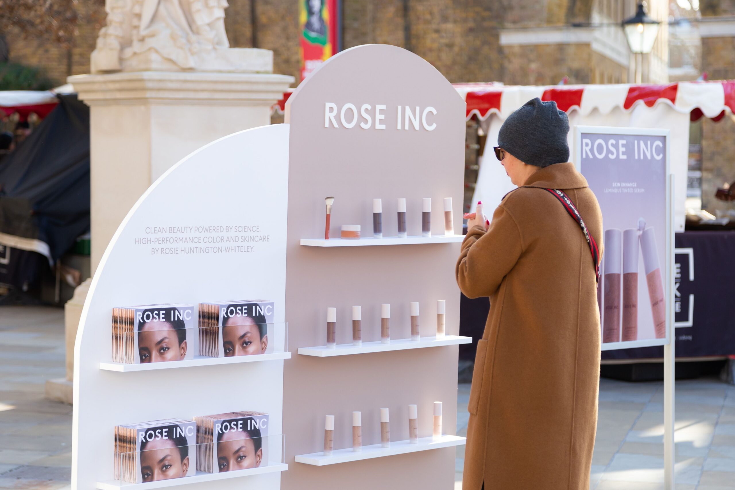 Person in brown coat and gray hat looks at cosmetics at a ROSE INC mobile pop up with shelves.