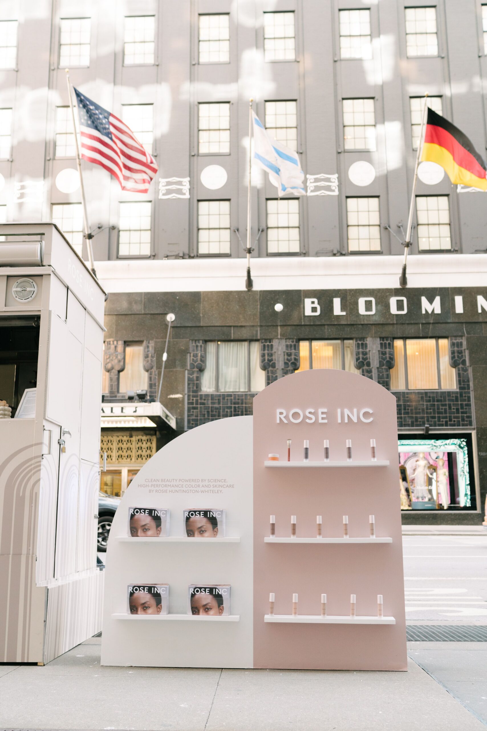 Rose Inc pop-up with beauty products on sidewalk; US, German, and another flag above Bloomingdale’s.
