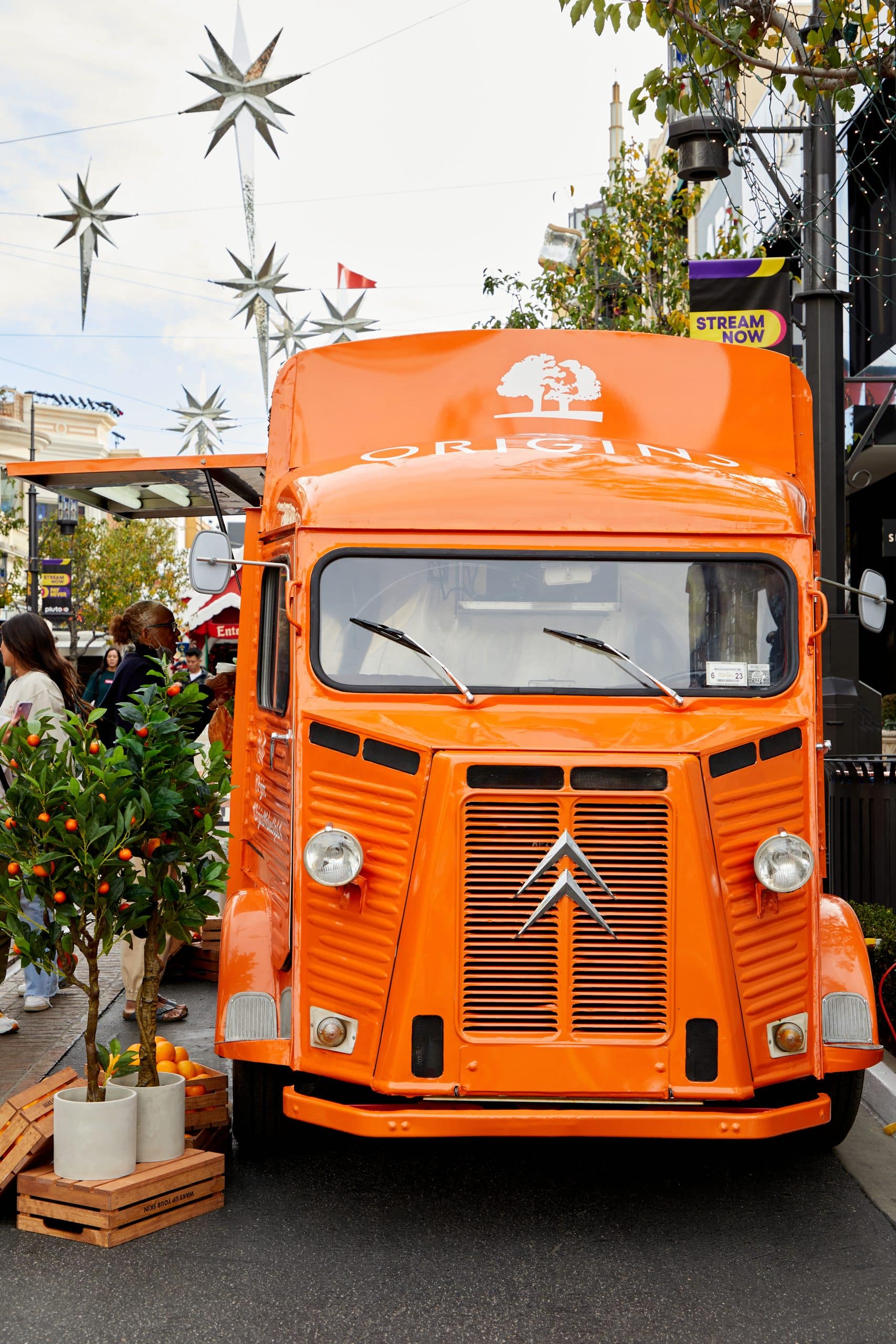 Orange vintage Citro&euml;n food truck on street with potted plants, people, and star-shaped lights.