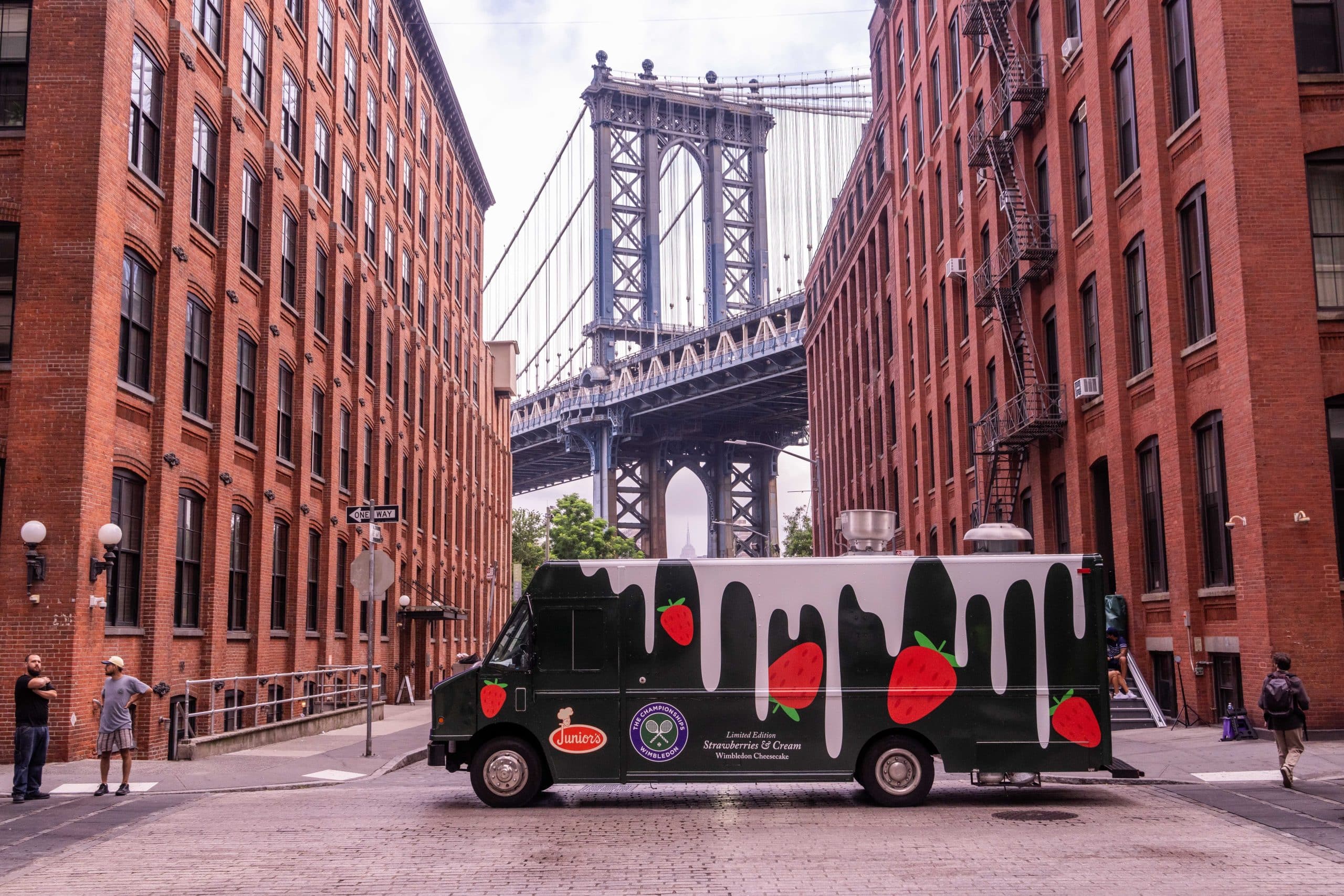A black, branded food truck with a watermelon pattern is parked on a cobblestone street between red brick buildings, with the Manhattan Bridge visible in the background in Brooklyn, New York.