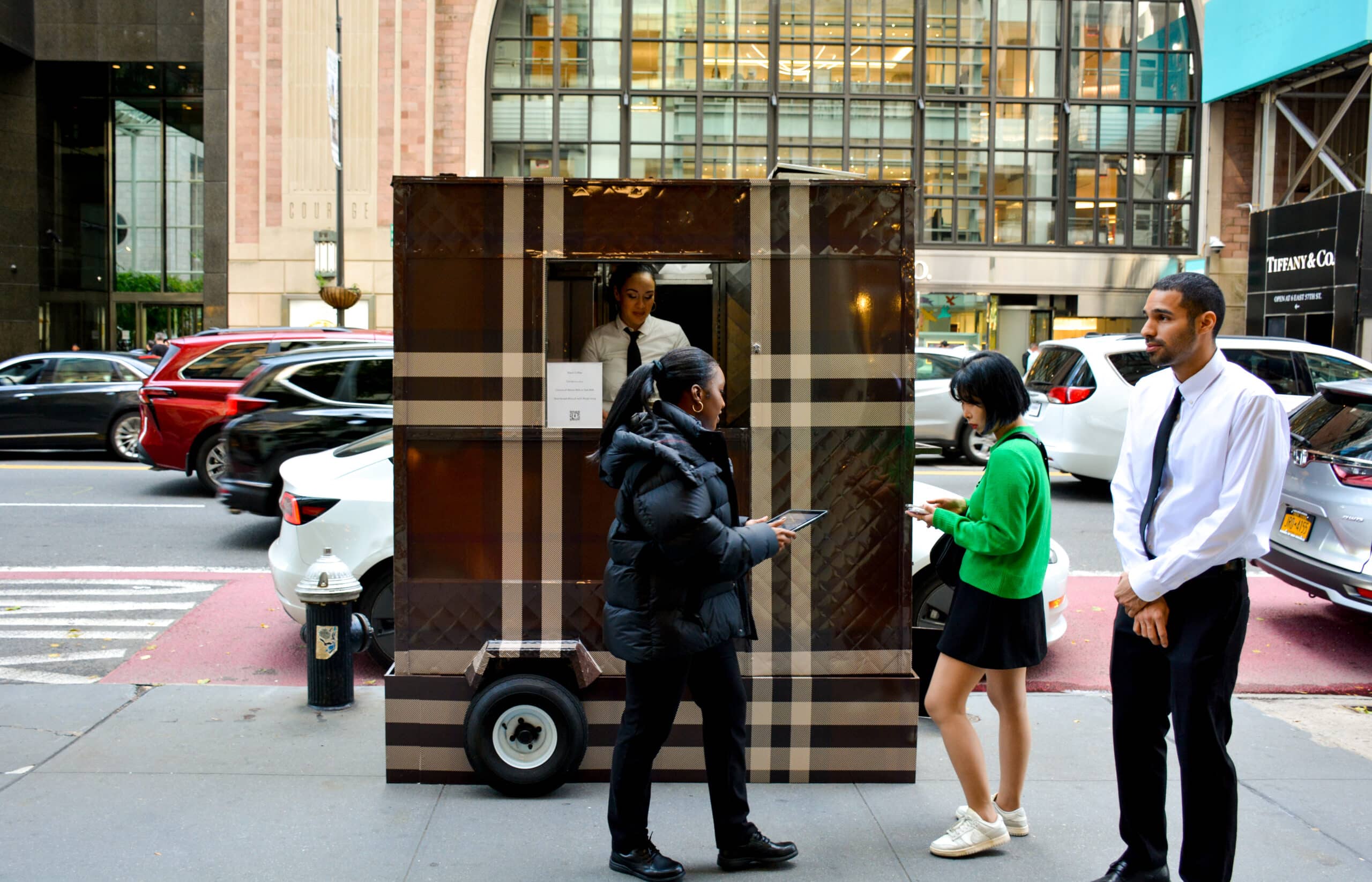 Two women use devices by a plaid street kiosk, with a man nearby and city buildings behind.